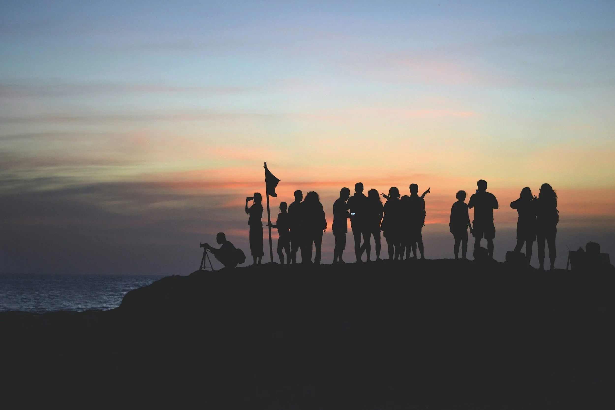 Silhouettes of people standing on a hill at sunset, some holding a flag, with a person taking a photo and others pointing or talking, against a colorful sky over the ocean.