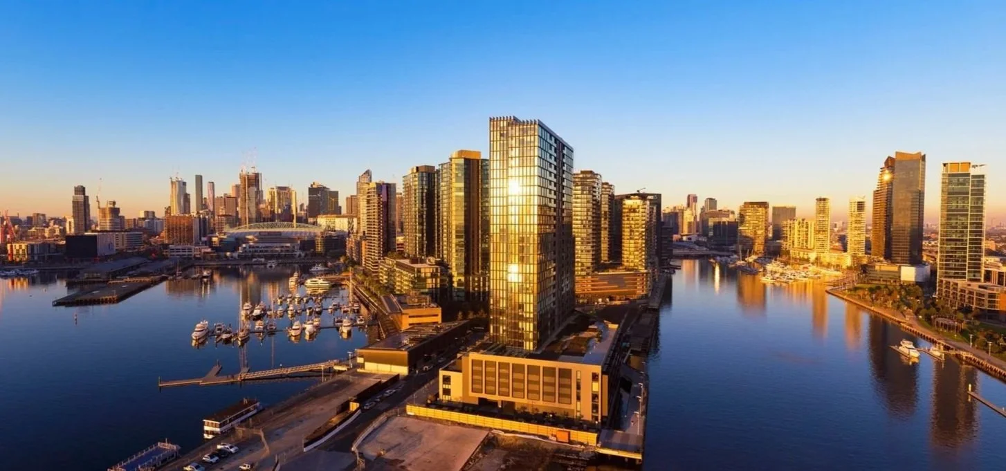 Skyline of a modern city with tall buildings and a river reflecting sunlight, viewed during sunset.