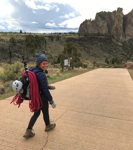 Eliza walking along a scenic path in Bend, Oregon, carrying climbing gear during an AdventurUs outdoor adventure weekend.