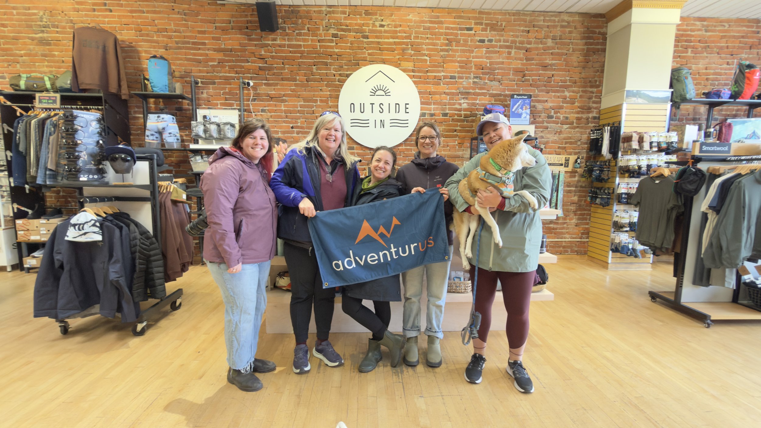 5 smiling women holding AdventurUs banner in Outside In store.