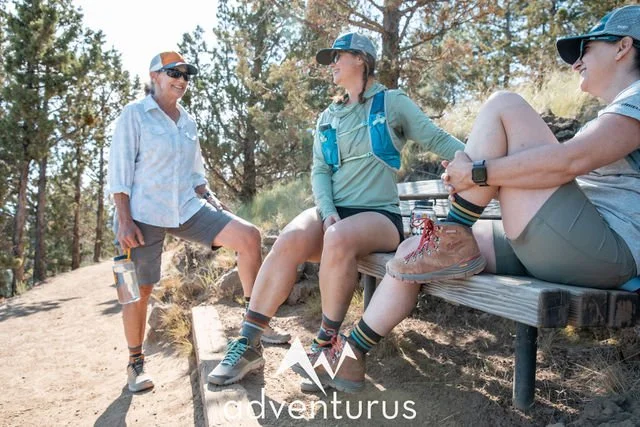 Three AdventurUs hikers chatting on a bench during a sunny trail break.