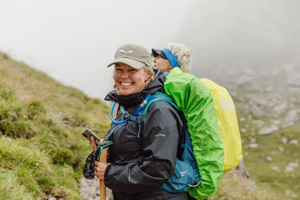 A smiling woman wearing a cap and a hiking jacket stands on a foggy trail during a trip organized by AdventurUs.