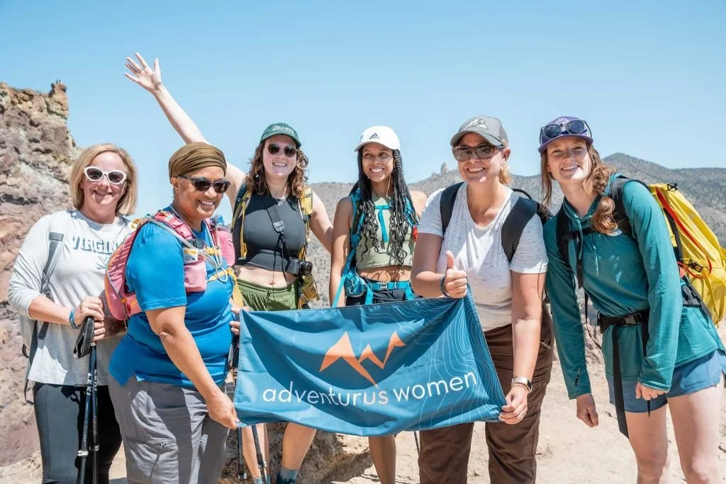 A group of women posing for an image with the AdventurUs banner on top of a mountain.