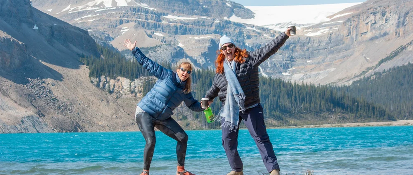 Two women, on an AdventurUs group trip, standing at a lake in the Canadian Rockies with mountains in the background, smiling and waving, dressed in outdoor winter clothing.