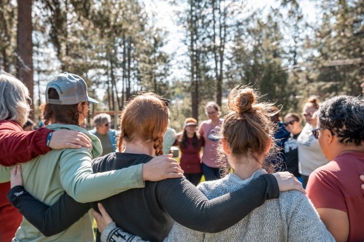 AdventurUs group standing arm in arm in a forest circle during an outdoor gathering.