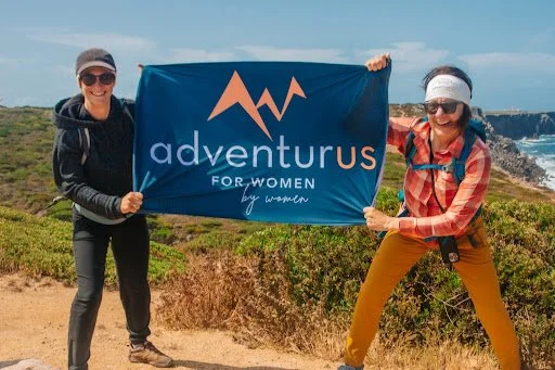 An image of Penny Bordokas during a hike holding the AdventurUs flag with another travel partner.