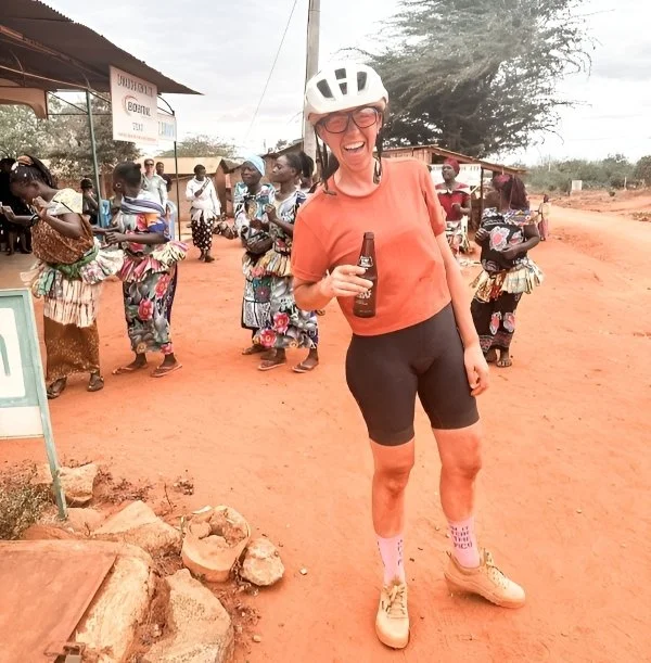 Jen Gurecki smiling during a cycling adventure in Africa, wearing a helmet and standing with local women in a village.