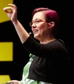 A white woman with glasses and short pink hair stands on a stage with a small microphone, smiling and speaking while gesturing with her left hand.