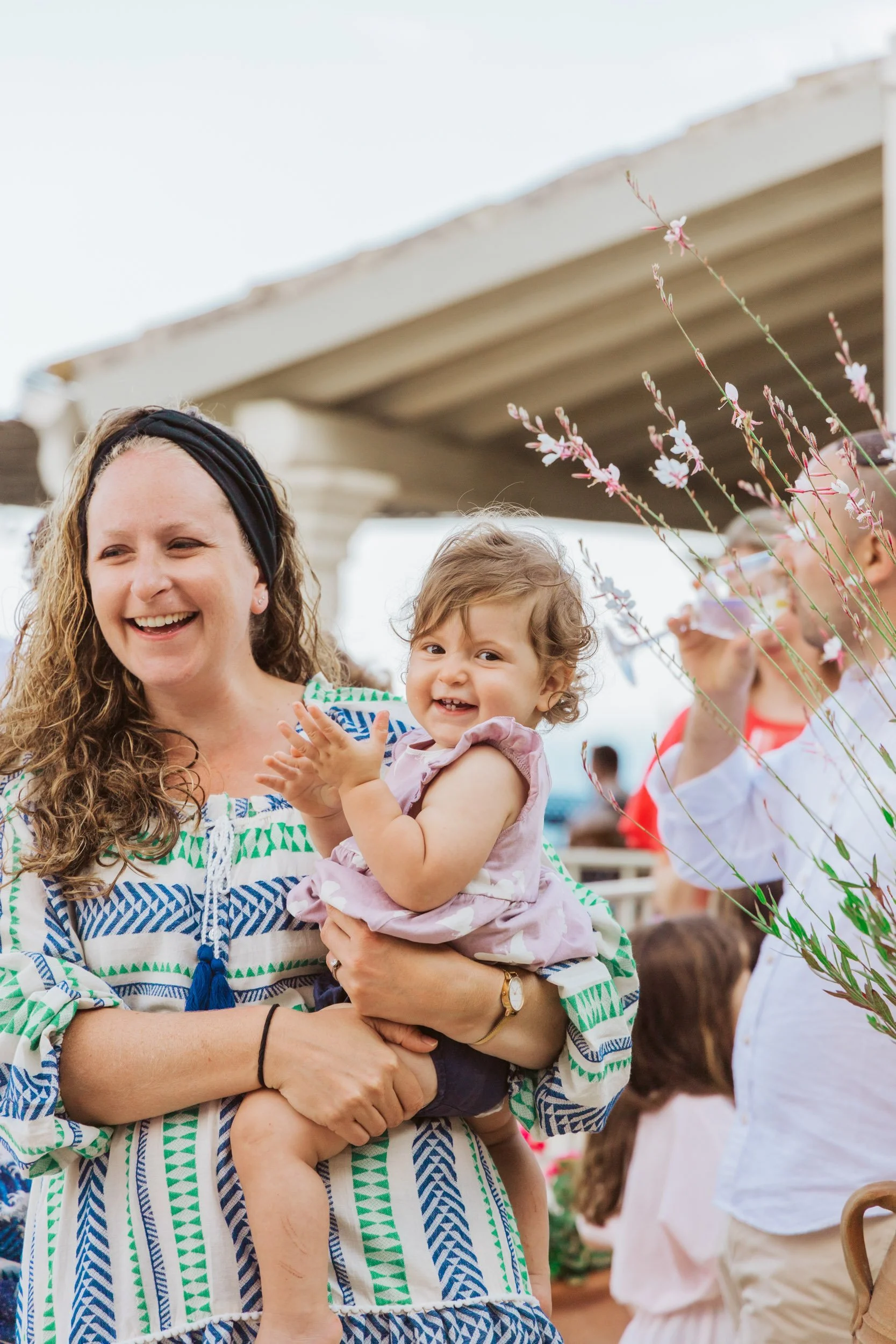 A woman with curly hair wearing a patterned dress and headband is holding a smiling young girl with curly hair in her arms. They are outdoors near a structure with a roof, and other people are visible in the background.