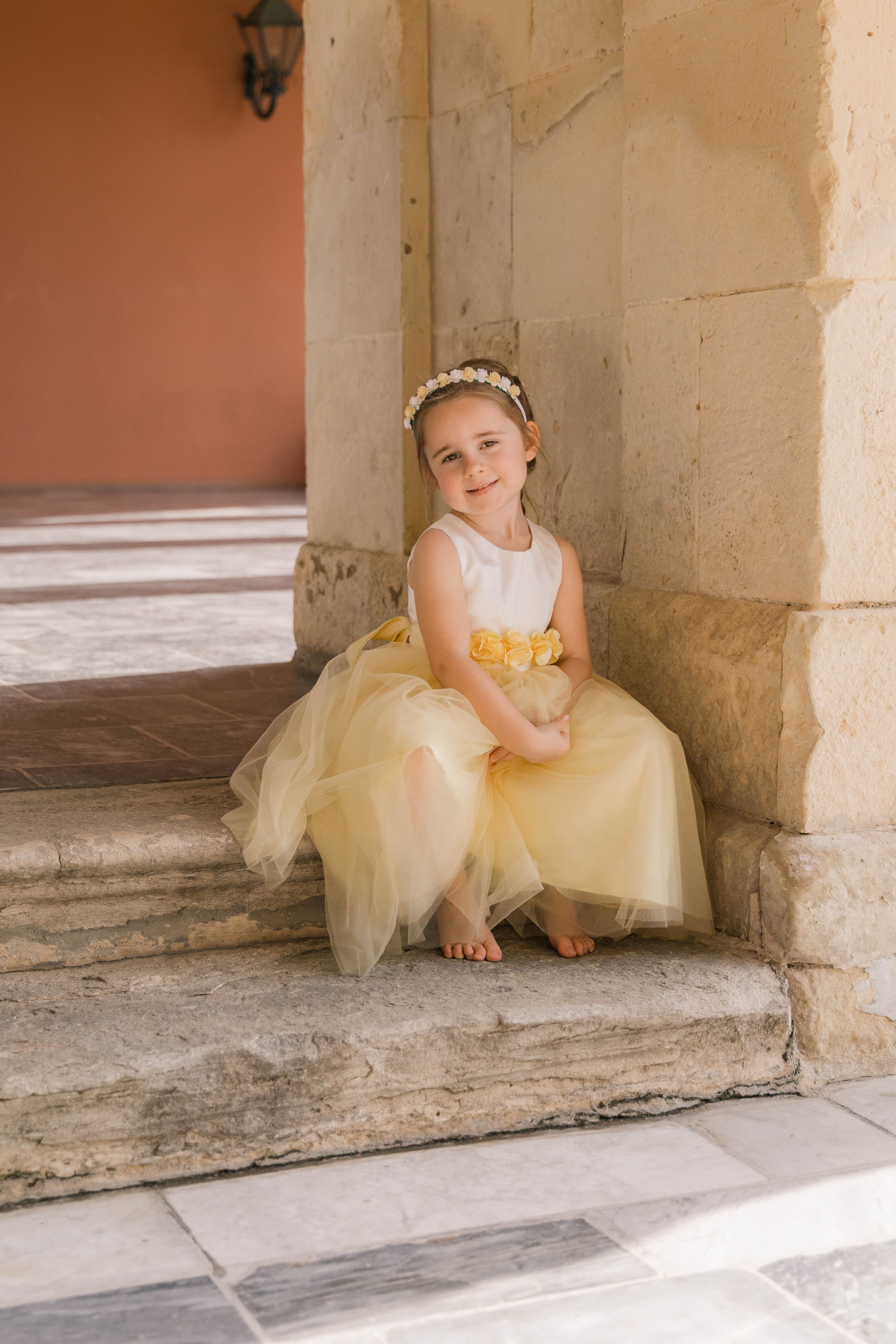 A young girl in a yellow dress and floral headband sitting on stone steps beside a stone wall.