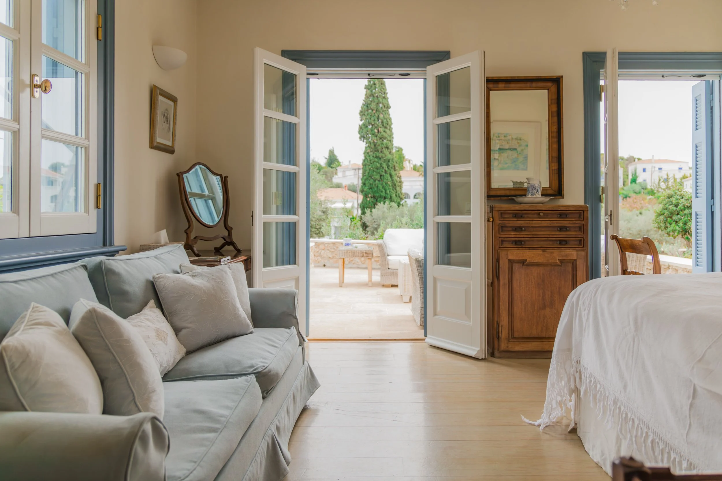 Spacious bedroom with open French doors leading to a balcony, featuring a bed with white bedding, a light-colored sofa with pillows, a wooden dresser with a mirror, and views of greenery and trees outside.
