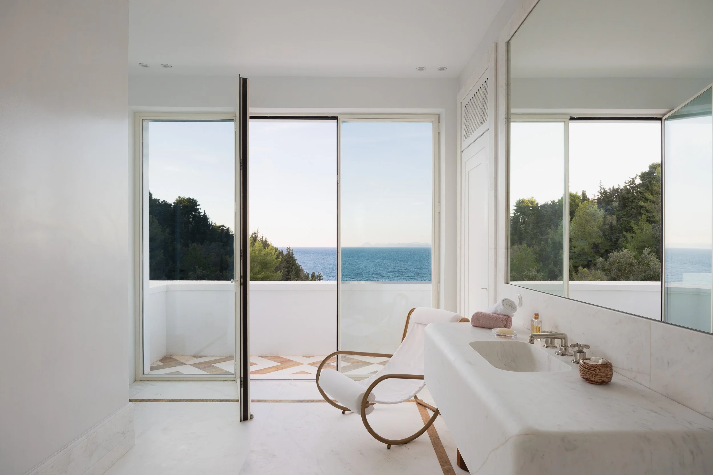 Bathroom with large mirror, white marble sink, beige towels, and a chair in front of a glass door leading to a balcony with ocean view and trees.