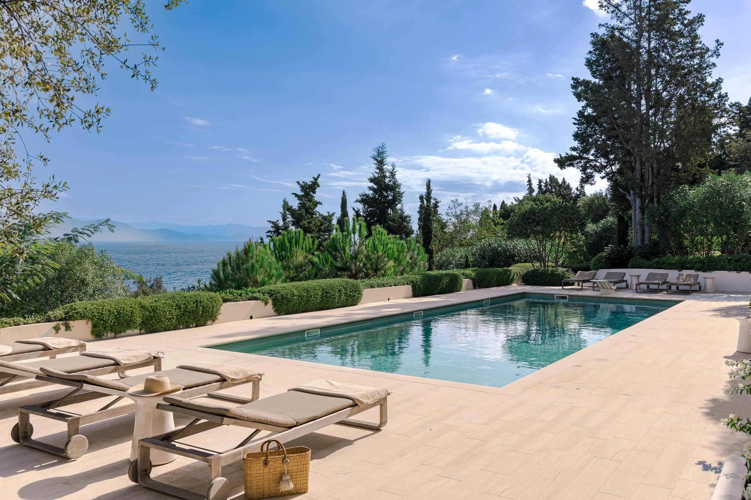 Swimming pool surrounded by lounge chairs, lush greenery, and trees with a view of the ocean and mountains in the distance on a clear sunny day.