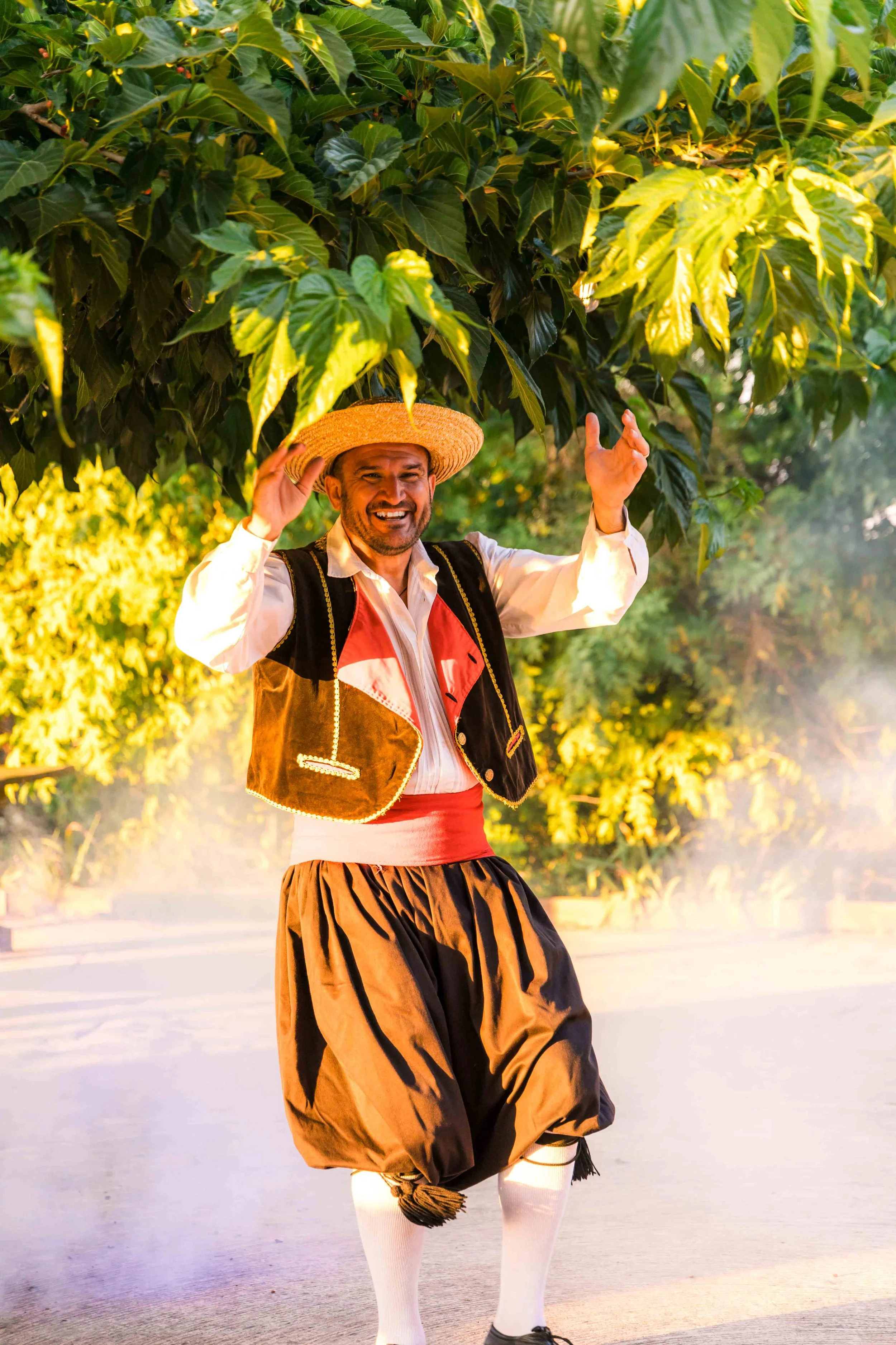 Man in traditional dress standing under a large tree, smiling, wearing a straw hat, vest, loose brown pants, and white stockings.