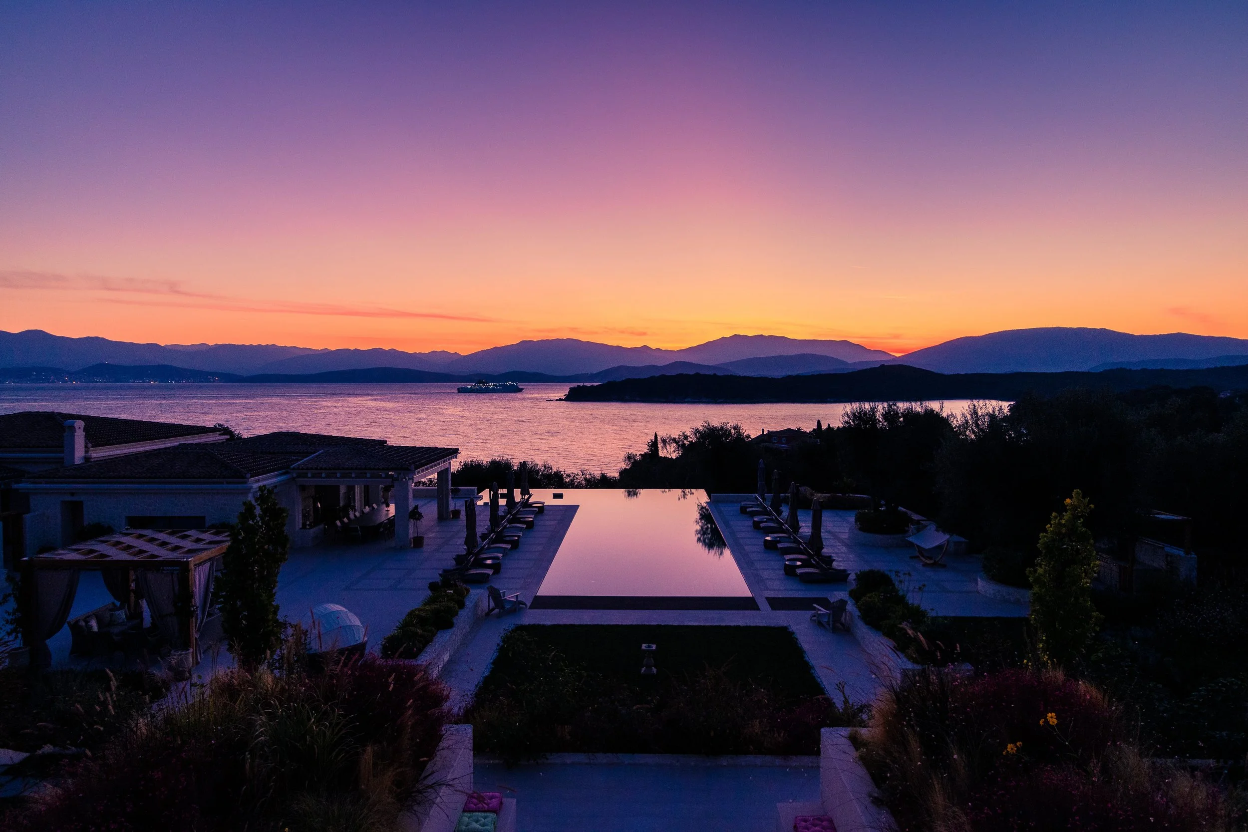 A luxurious outdoor pool area overlooking a lake and mountains at sunset, with lounge chairs, umbrellas, and lush landscaping.