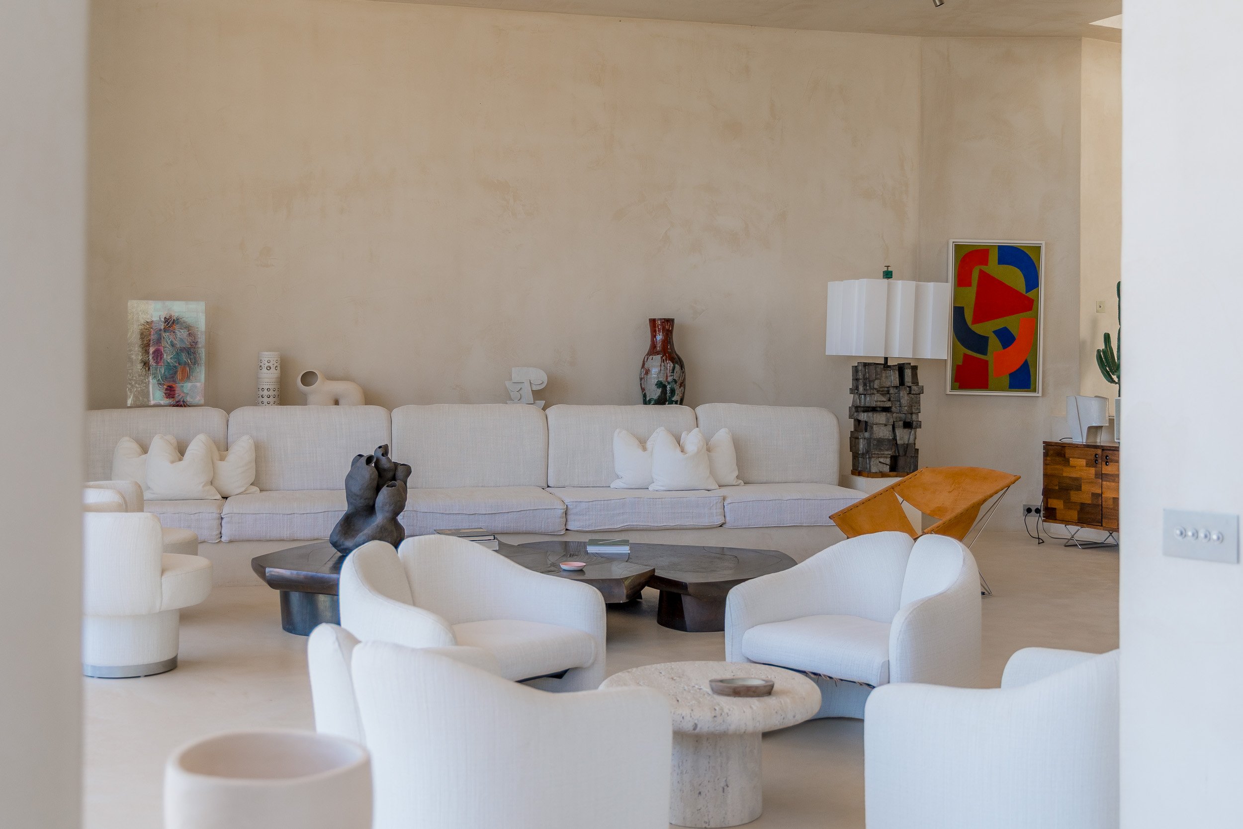 A spacious living room with white contemporary furniture, including armchairs, a large sectional sofa with pillows, and unique artworks on the walls. Decor includes a dark wooden coffee table, a sculptural piece, a large abstract painting, and a wood