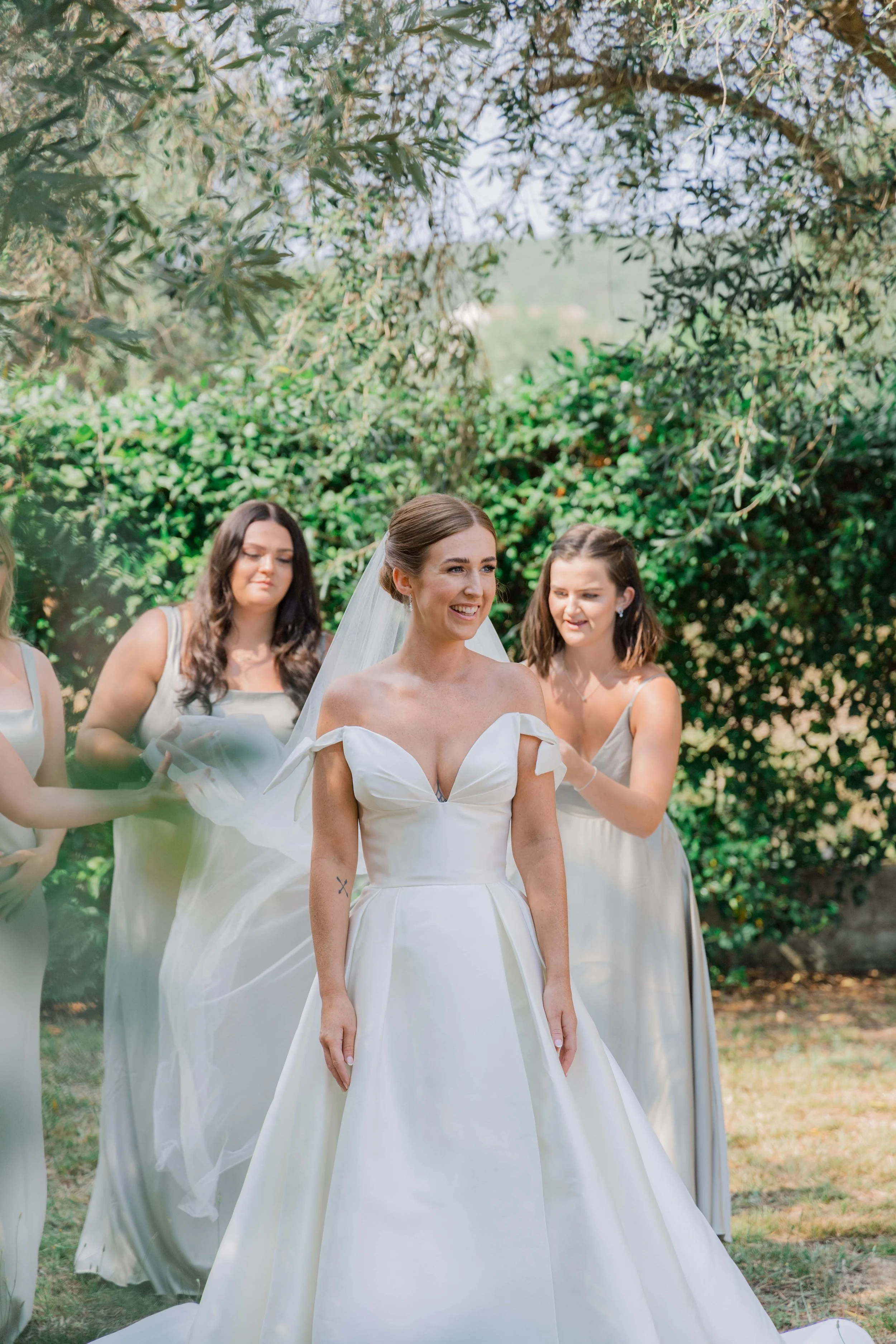 Bride in a white wedding gown smiling as her bridesmaids help her prepare outside