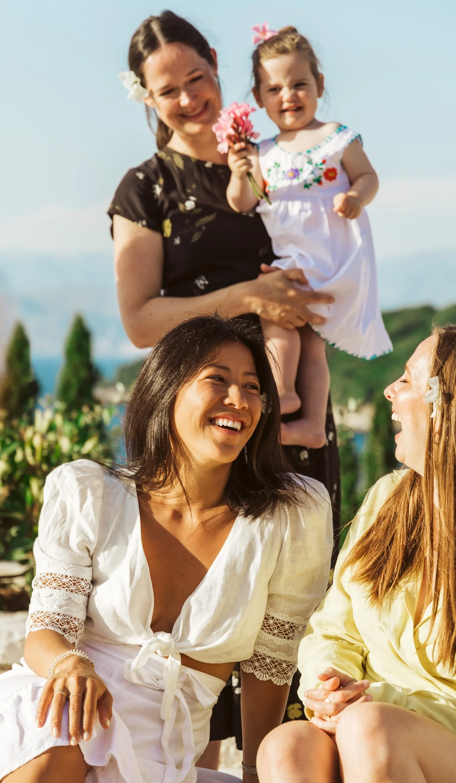 Four women and a girl are outdoors, enjoying a sunny day. The woman on the left is smiling and sitting, while the woman on the right is also smiling and sitting next to her. The woman standing in the background is holding a young girl who is holding 