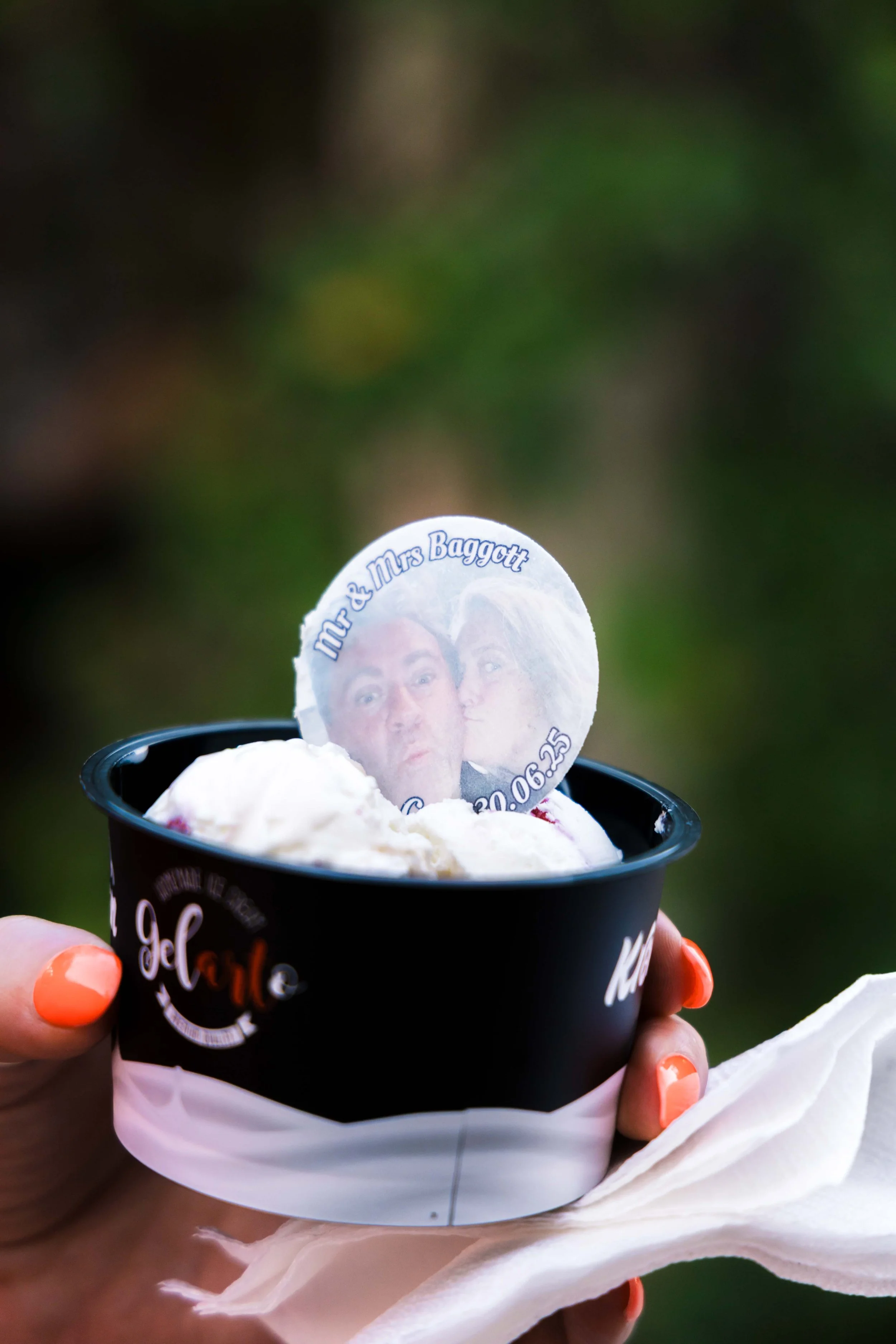 A person's hand with coral nail polish holding a black cup of ice cream, topped with a photo souvenir of a couple and the text 'Mr & Mrs Baggott' and a date, against a blurred green outdoor background.