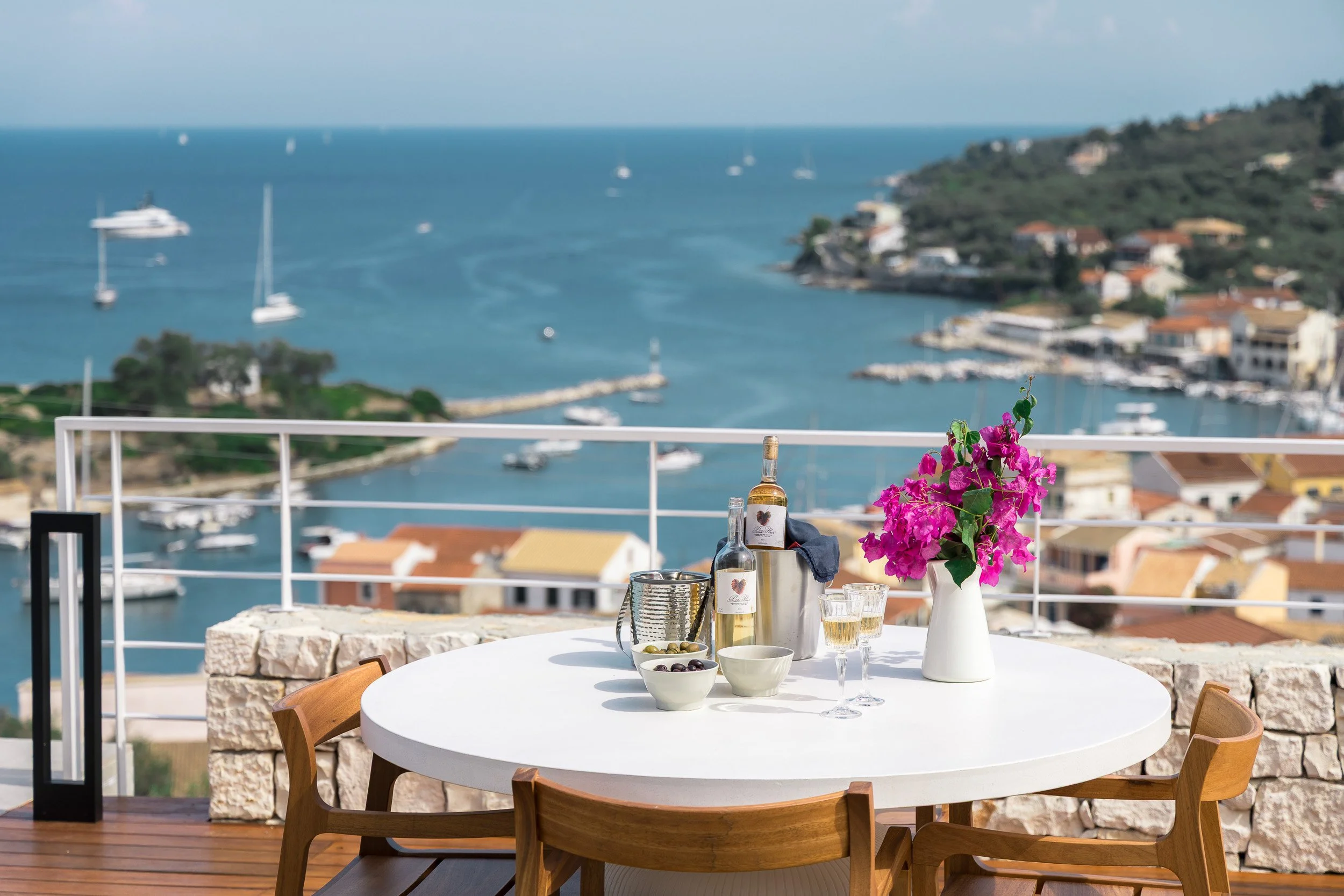 A round white table with a pink flower vase, two bottles of wine or spirits, glasses of white wine, bowls of olives, and a pitcher on a balcony with a view of a harbor with boats and houses.