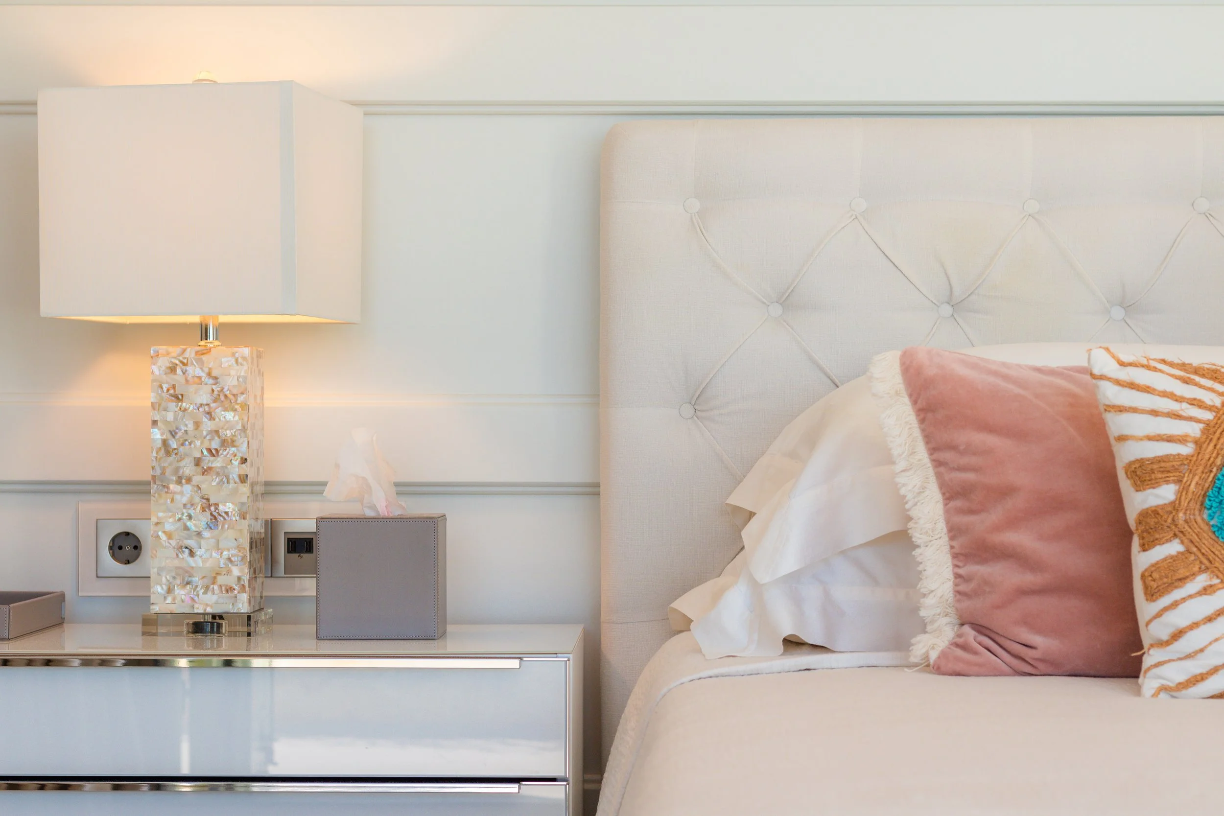 Close-up of a bedroom nightstand with a lamp, tissue box, and electrical outlets, next to a bed with white and pink pillows.