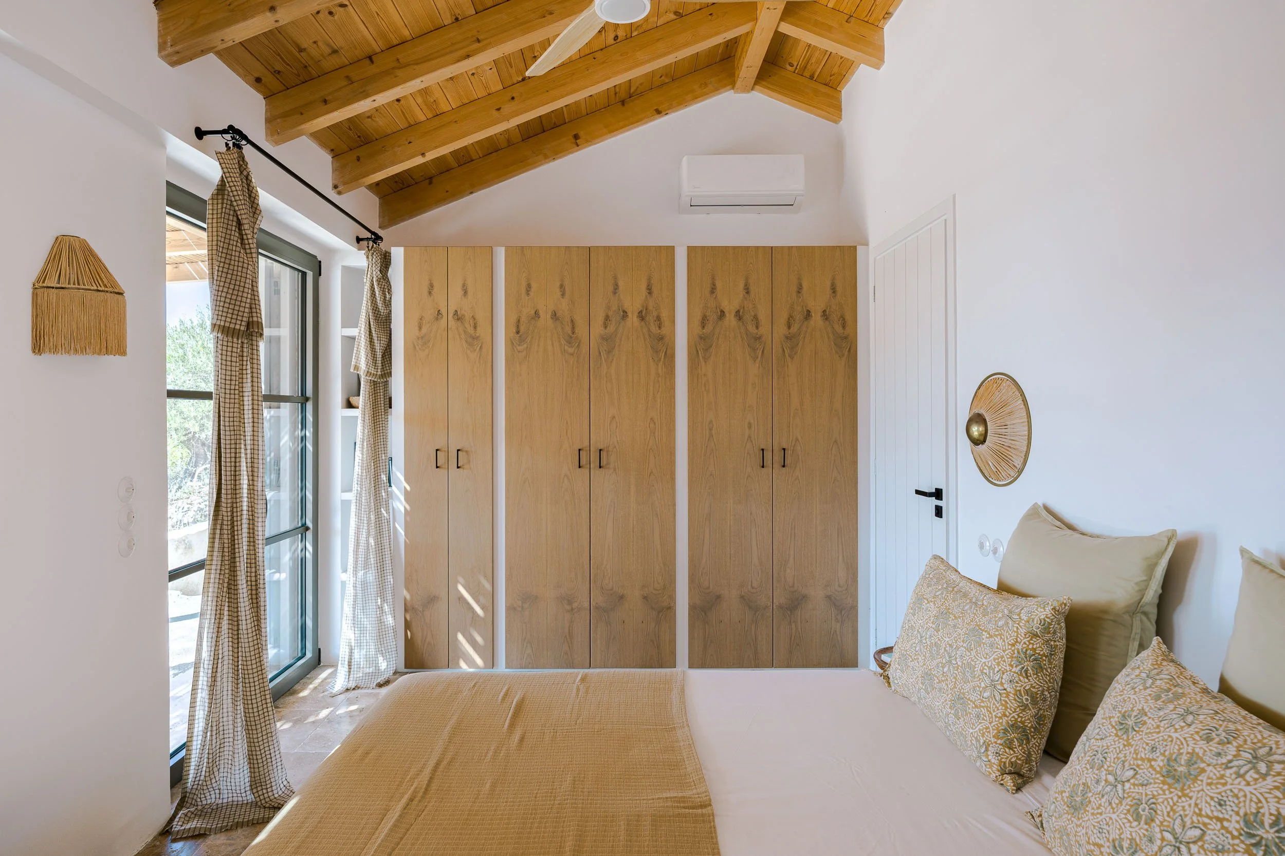 A bedroom with a wooden ceiling, beige bed with patterned pillows, wooden closet doors, sliding glass door with curtains, air conditioning unit, and decorative wall art.