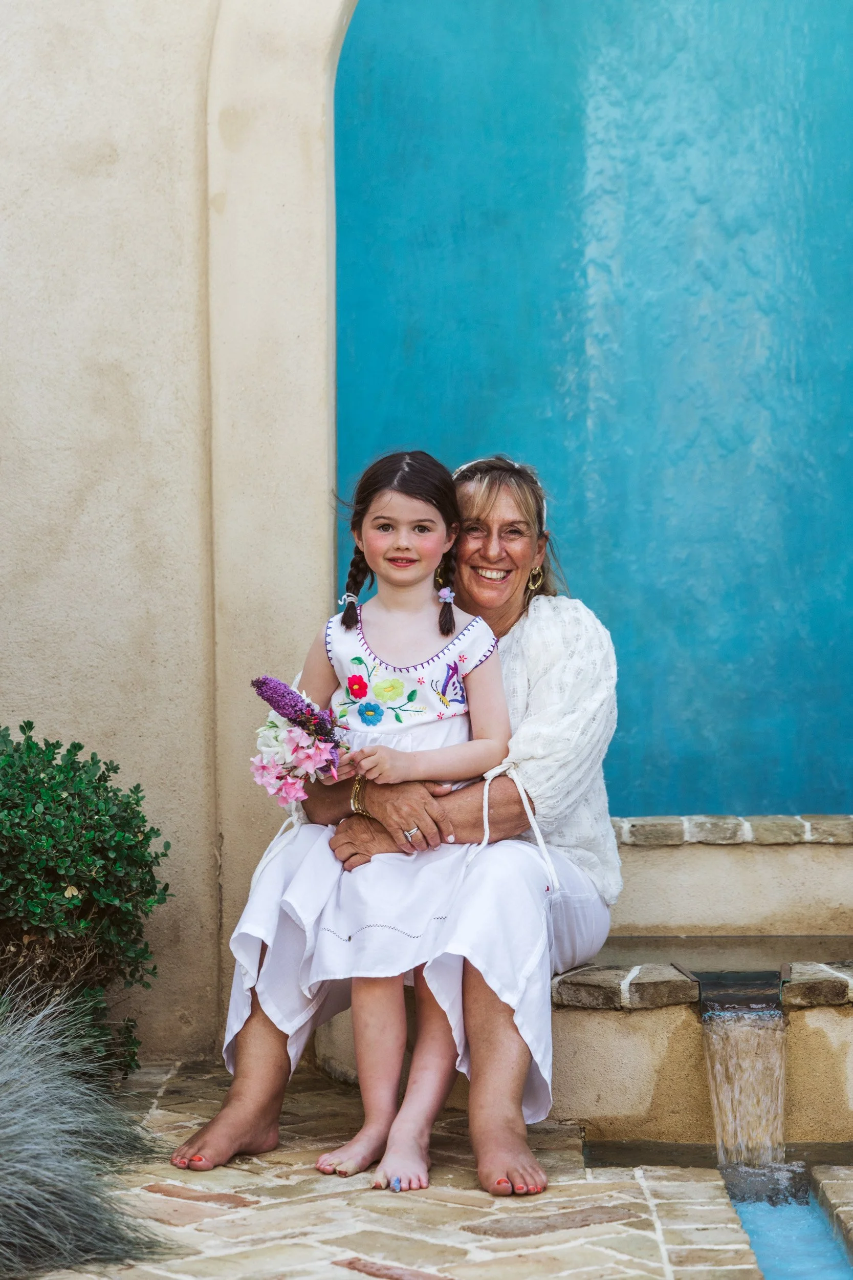 A young girl and an older woman sit together on a stone bench, smiling. The girl holds a bouquet of flowers. They are outdoors against a beige wall with a blue feature and a small water fountain.