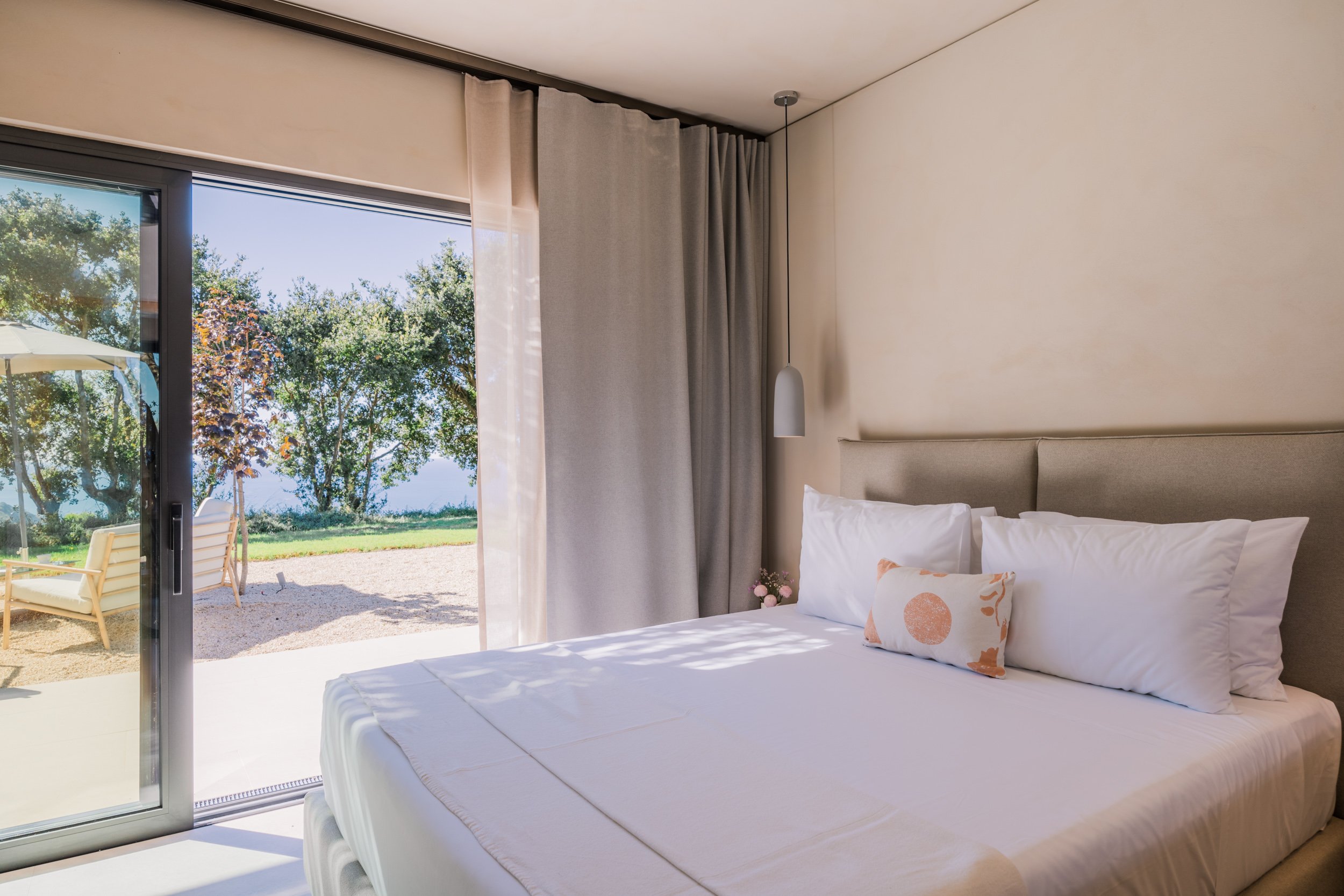 A bedroom with a bed, white sheets, and pillows near glass sliding door leading to an outdoor patio with trees and outdoor furniture.