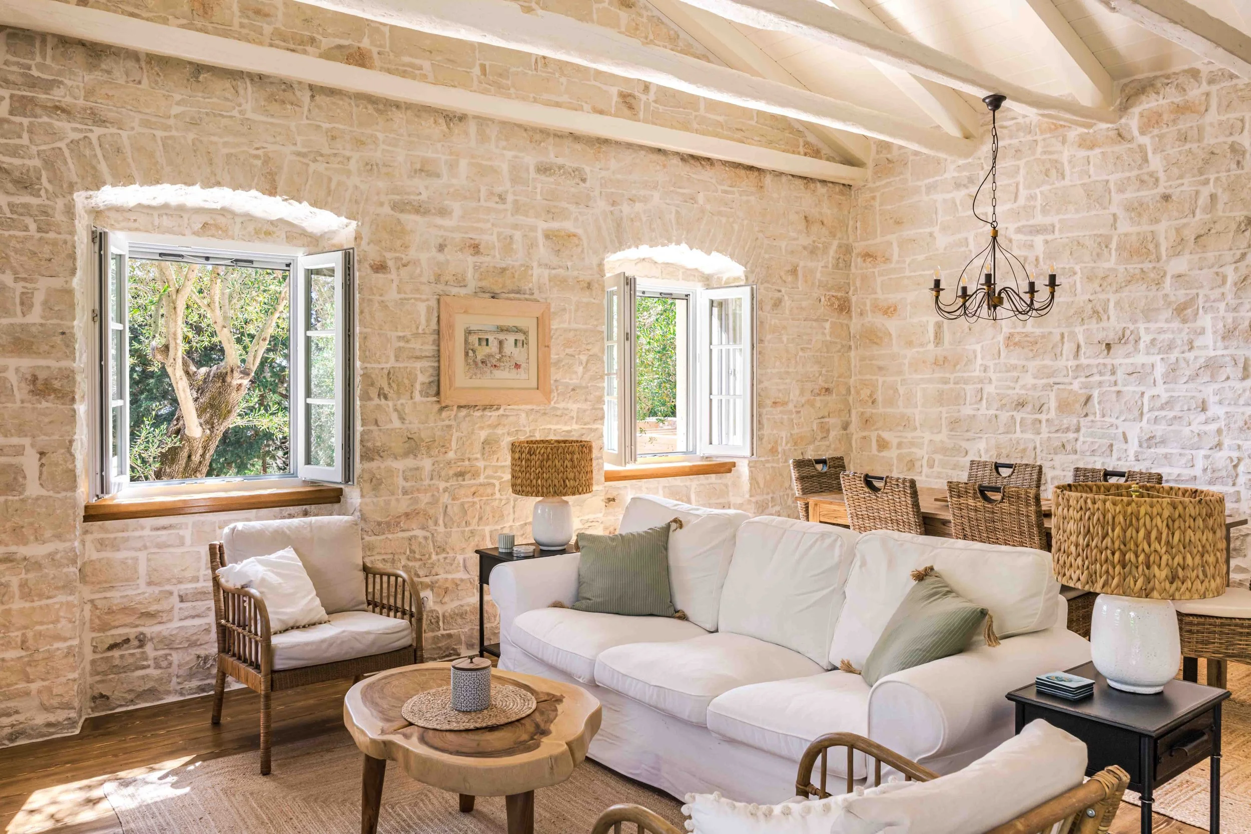 Living room with beige stone walls, wooden beams, two open windows, a white sofa with green pillows, a wood and wicker chair, a black side table, and a rustic wooden coffee table, decorated with lamps and framed artwork.