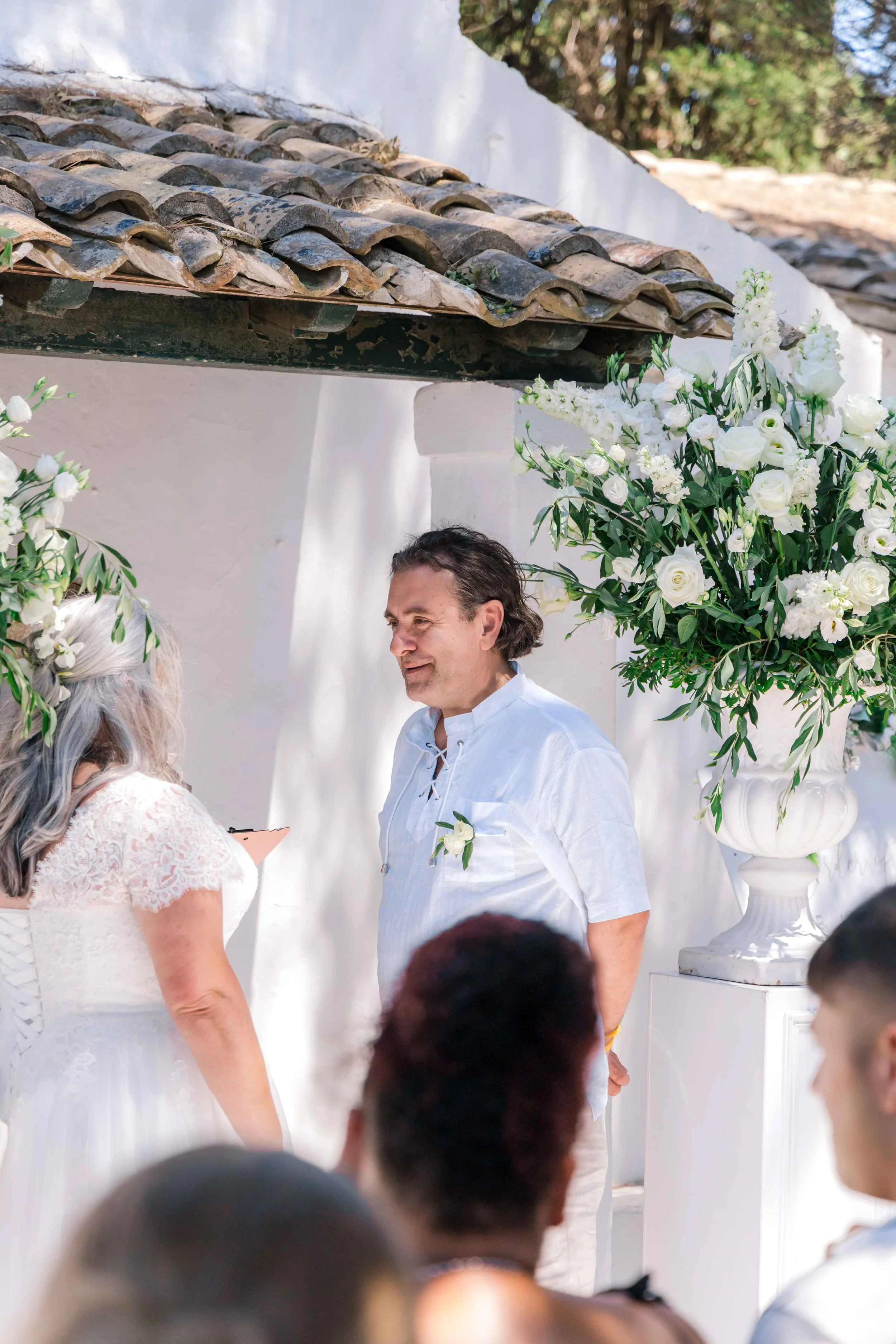 A couple gets married in a white outdoor setting with white floral arrangements, one large bouquet on a white pedestal, and white walls, during daytime.