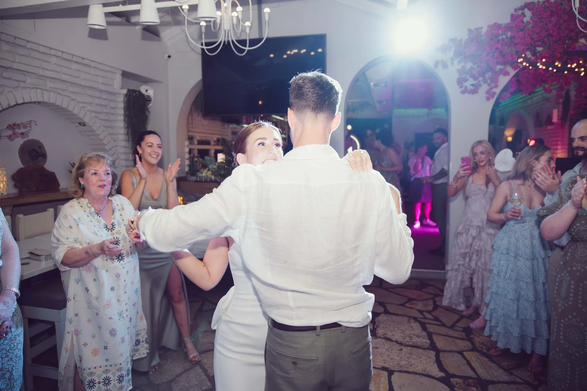 People dancing and celebrating at a wedding reception, with a bride and groom in the center dancing, surrounded by guests clapping and smiling indoors with decorated walls and ambient lighting.