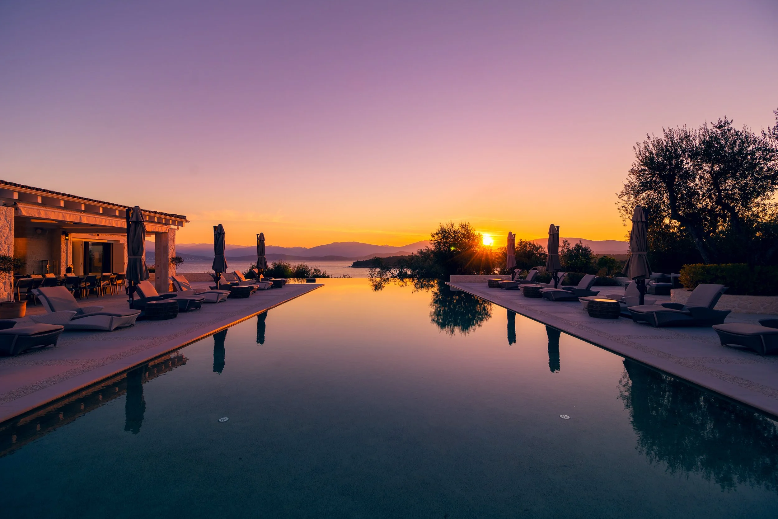 Sunset view over infinity pool at a luxury resort, with poolside lounge chairs and umbrellas on both sides, overlooking water and mountains in the distance.