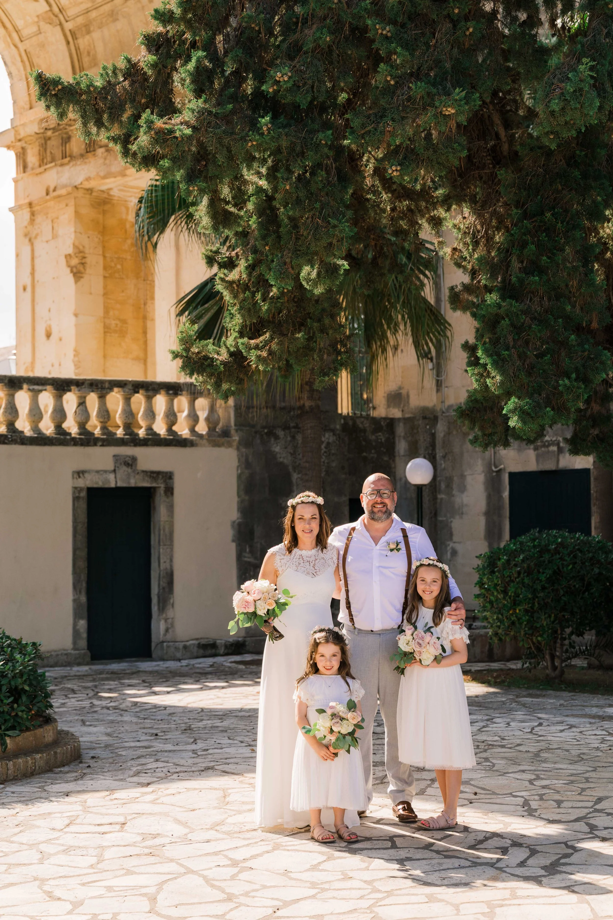 A wedding couple with two young girls stands outdoors on a sunny day, holding bouquets, in front of historic stone buildings and greenery.