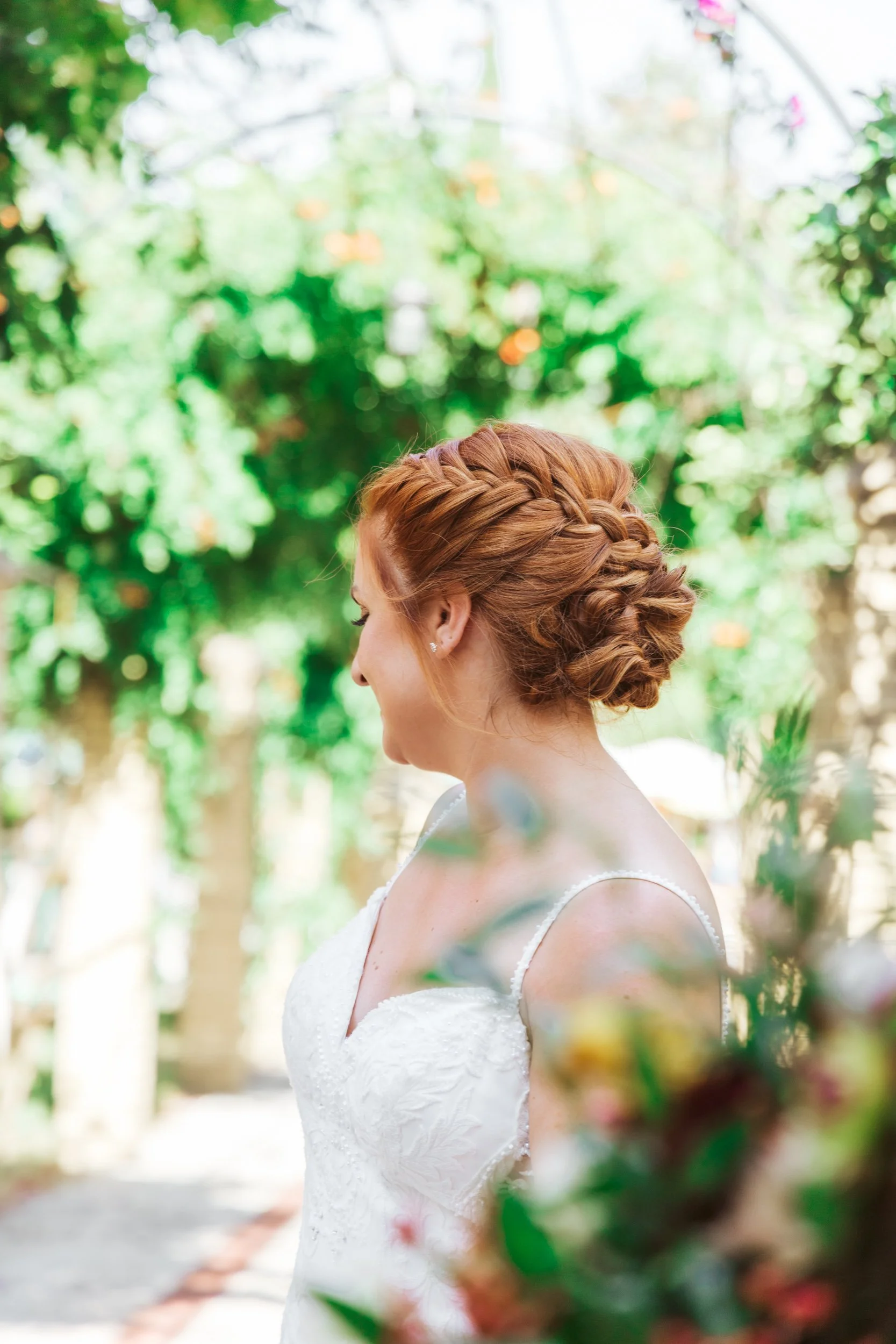 A bride with braided red hair in a white wedding dress standing outdoors among green foliage.
