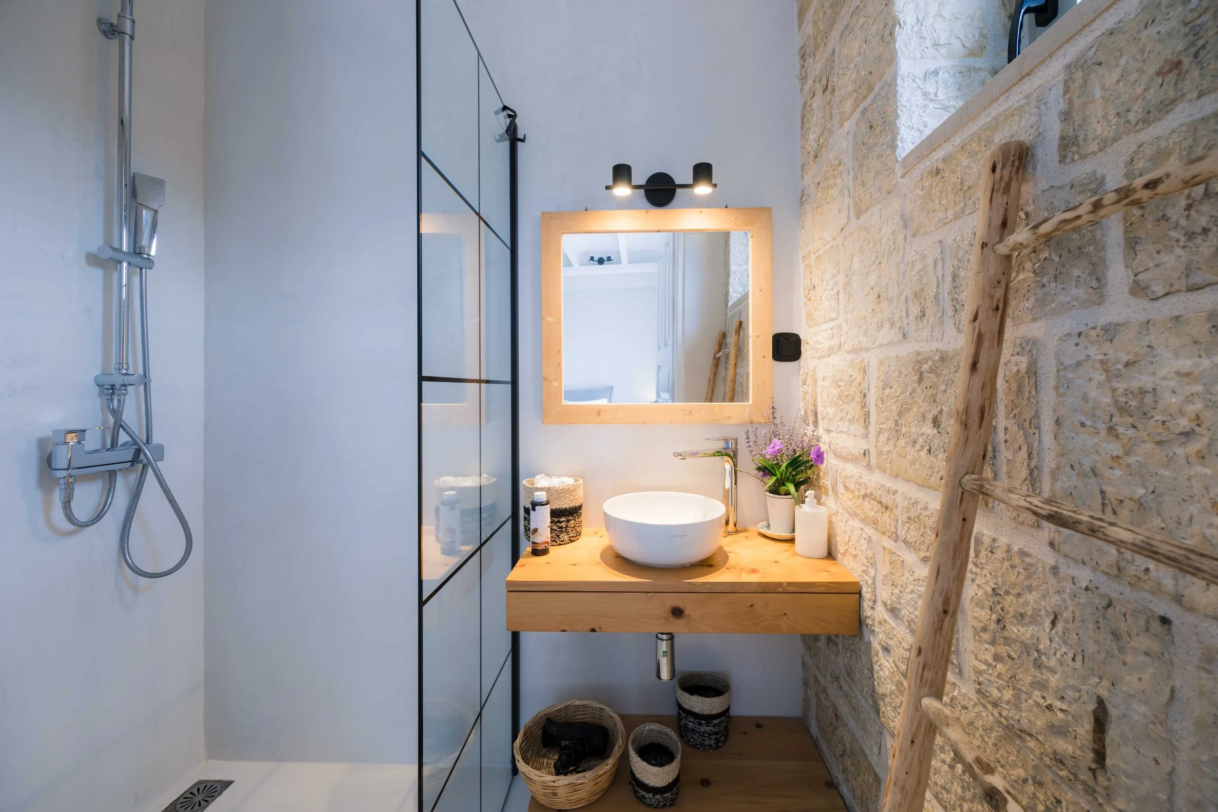 Bathroom with stone wall, wooden ladder, black and white decor, sink with mirror, shower area on the left.