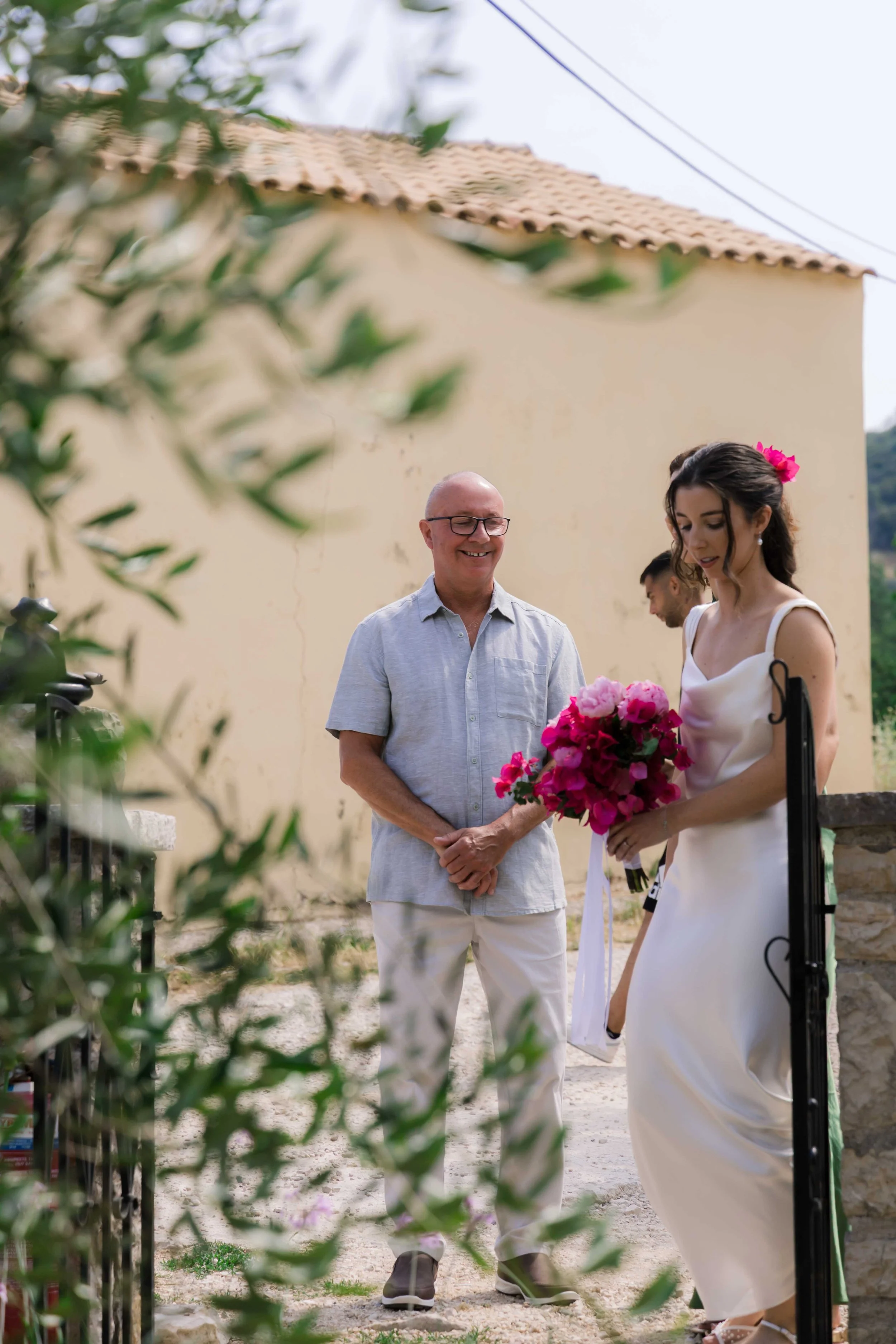 A woman in a white dress holding a bouquet of pink flowers stands outside near a gate, talking to a man in a light blue shirt and white pants, with a third person in the background, in a sunny outdoor setting.