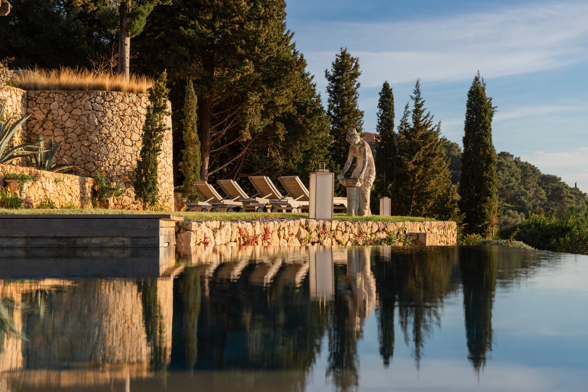 Luxury outdoor pool area with stone wall, lounge chairs, statues, and trees during sunset.