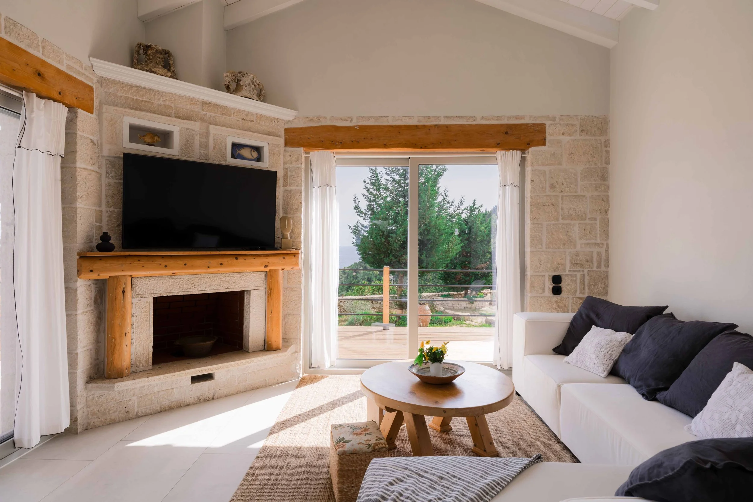 Living room with stone fireplace, wooden mantel, large sliding glass door leading to a balcony with trees, white curtains, black and white sofa, wooden coffee table with a bowl of flowers, and a small cushioned stool.