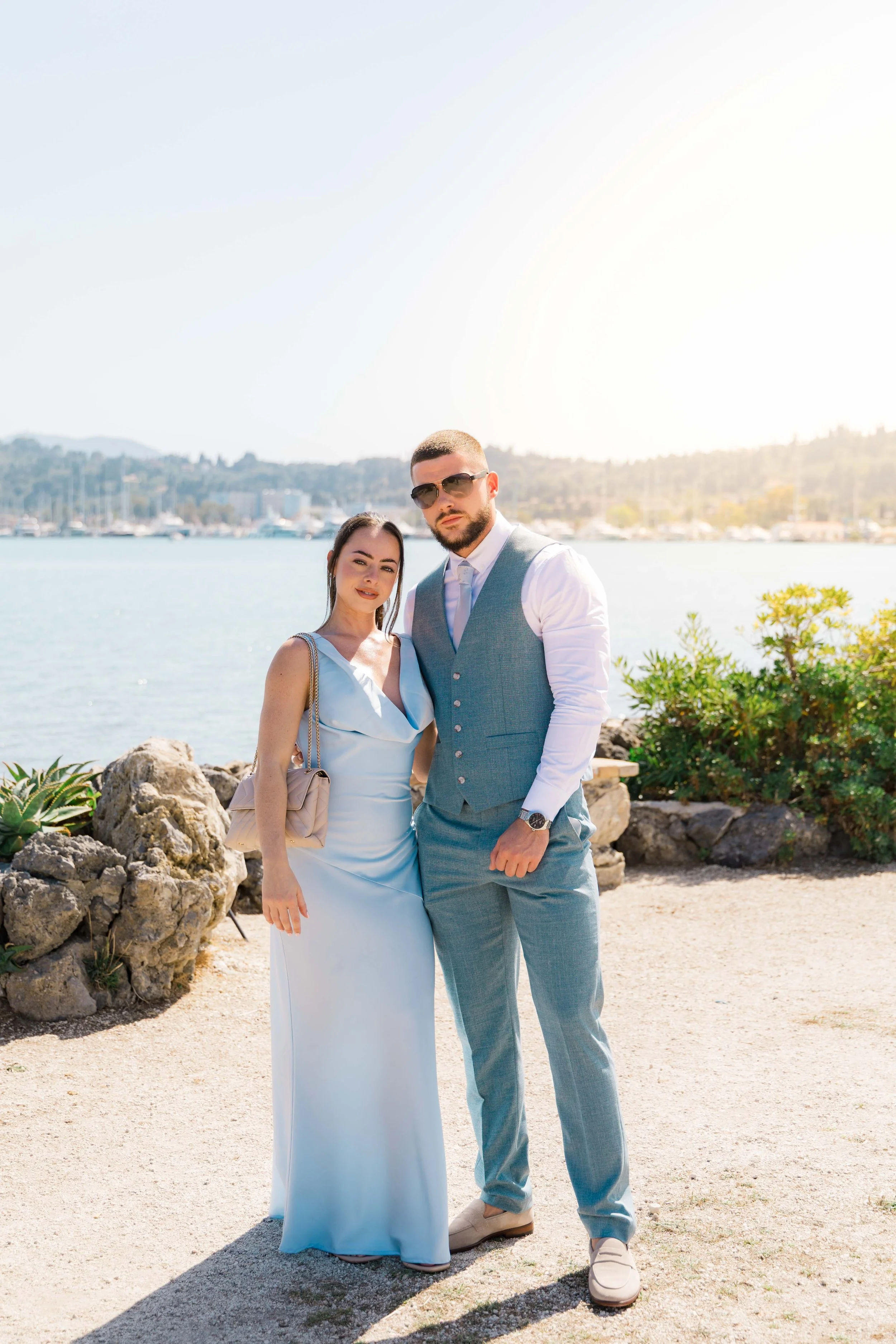A man and woman dressed in formal attire standing outdoors near water, with boats and hills in the background, on a sunny day.