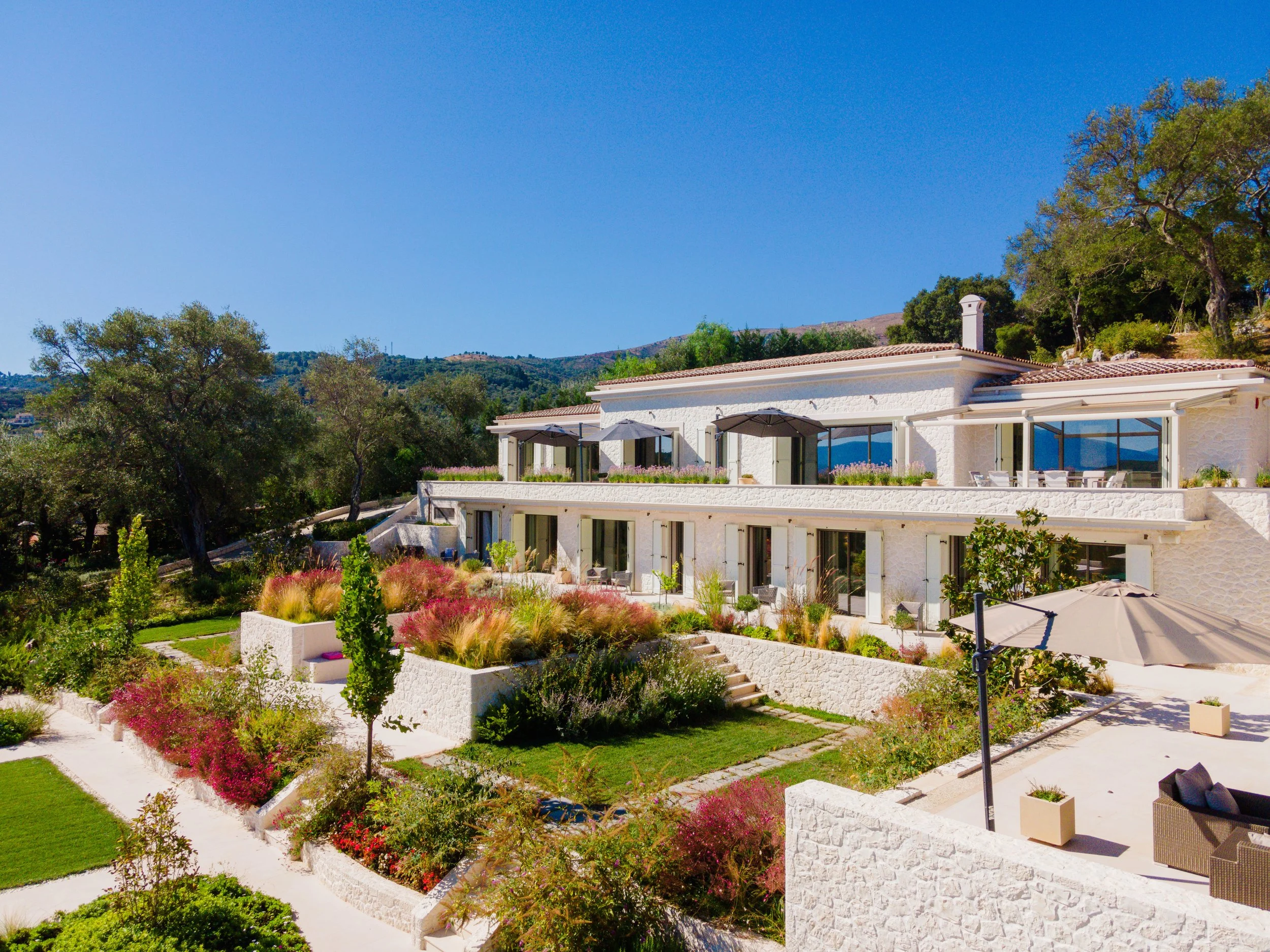 A modern white multi-story house with large windows, surrounded by a lush garden with colorful plants and trees, on a sunny day with a clear blue sky.