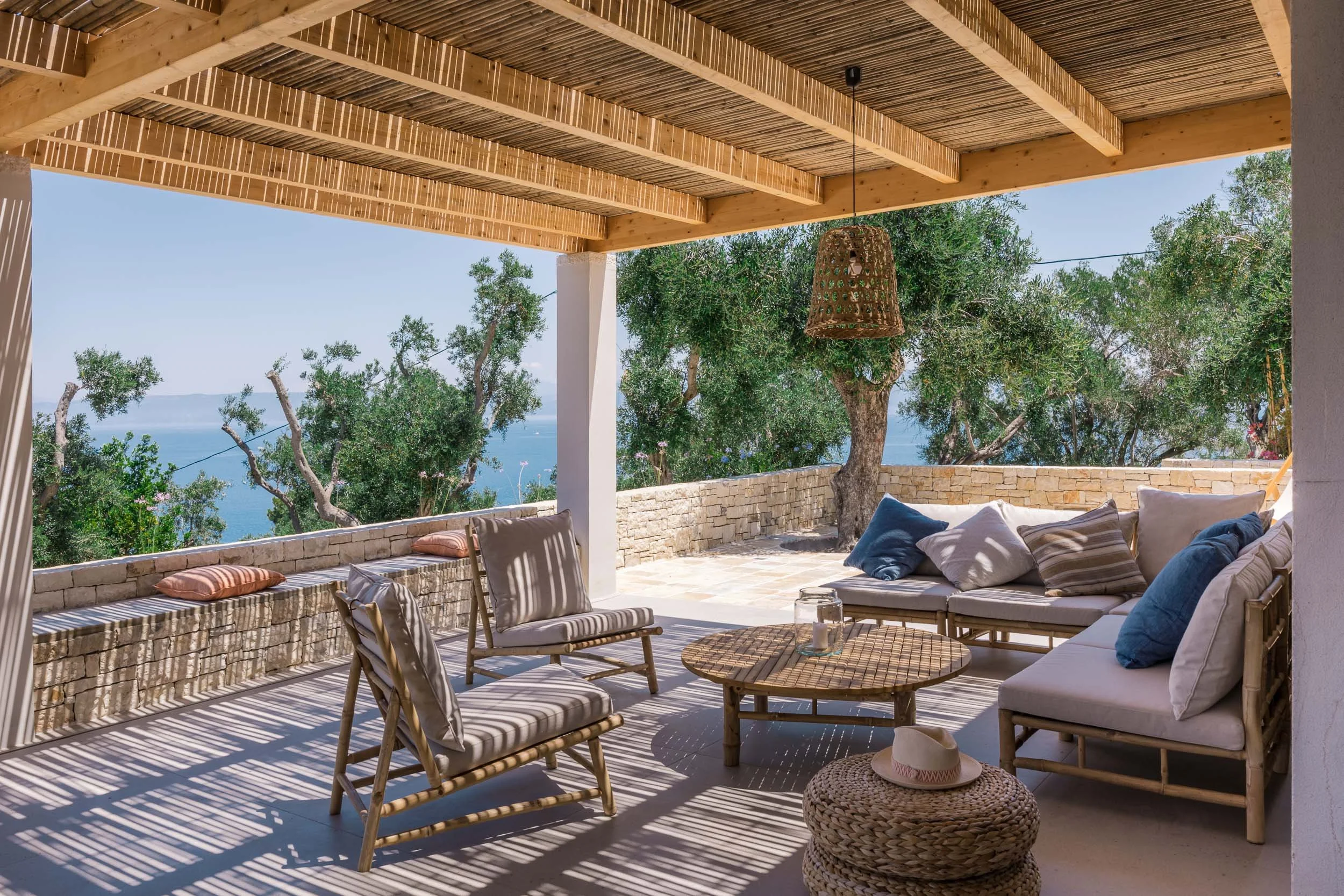 Outdoor patio with seating area including a white cushioned sofa, chairs, a round coffee table, wicker ottoman, and a hanging wicker lamp, with trees and an ocean view in the background under a wooden pergola.