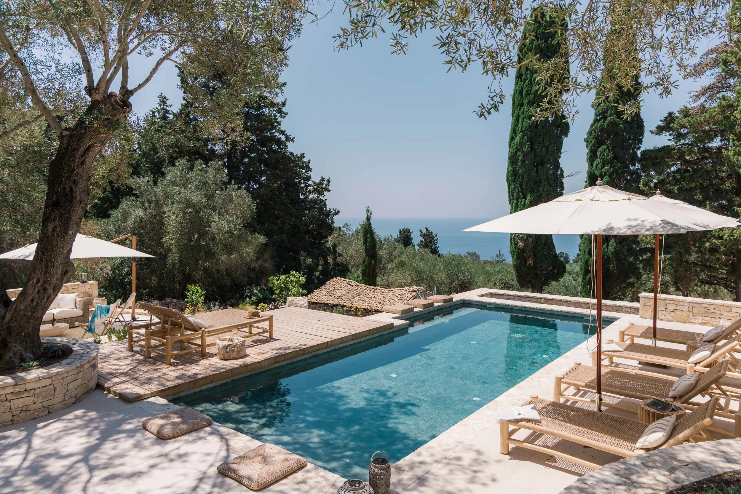 Luxury outdoor swimming pool with lounge chairs, umbrellas, surrounded by trees and greenery, with a view of the ocean in the distance.
