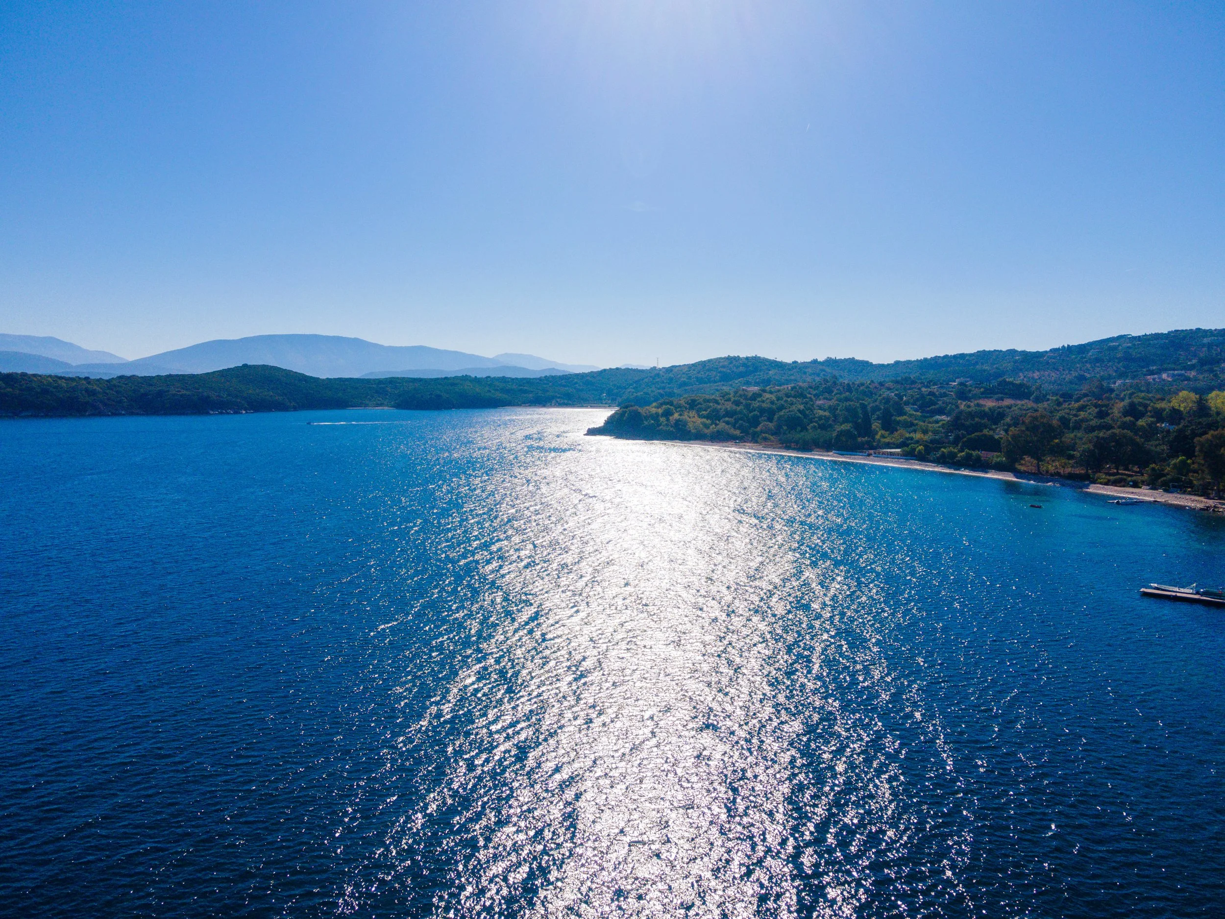 Aerial view of a large blue lake with shimmering water, bordered by green hills and mountains under a clear blue sky.