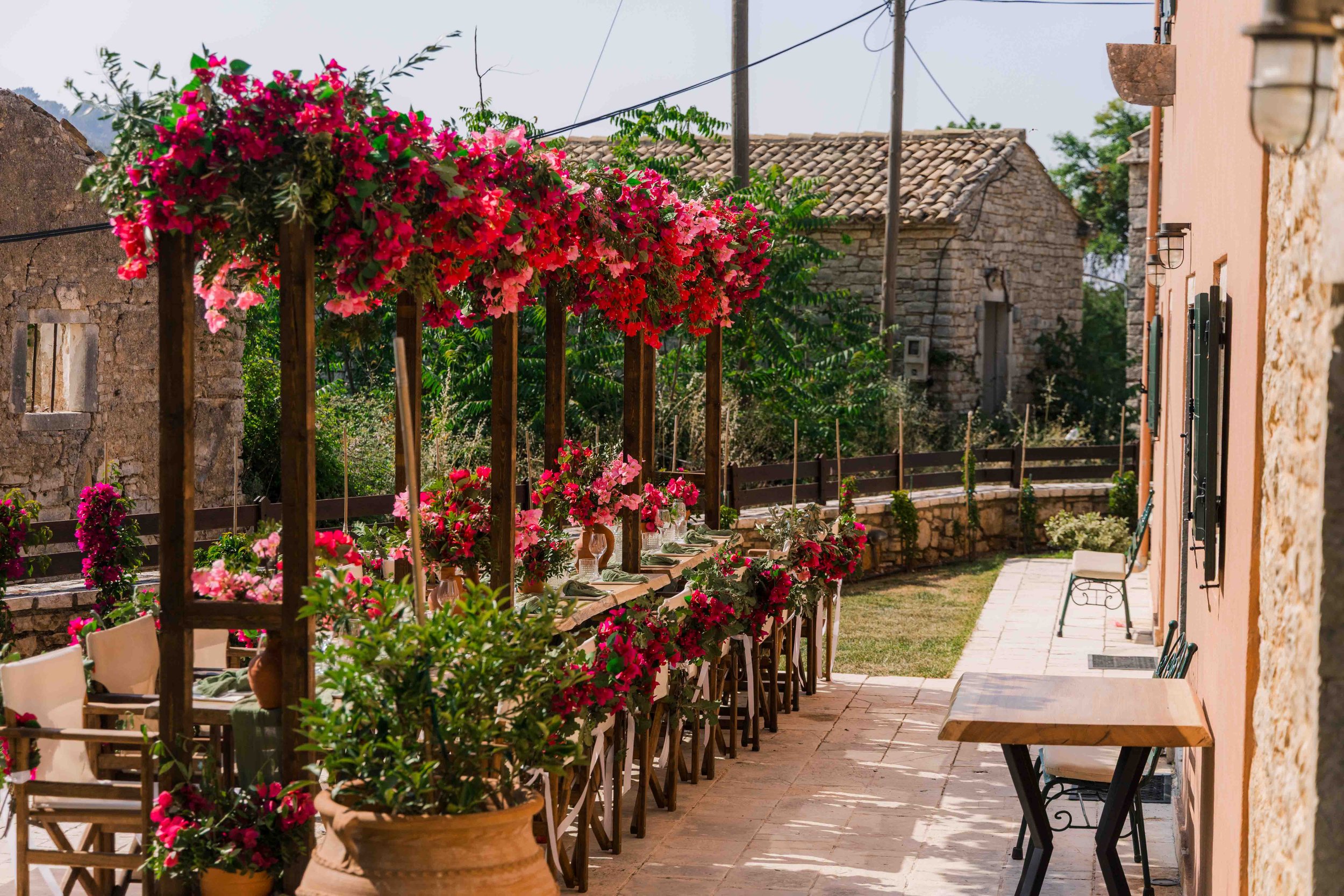 Outdoor patio with pink and red flowers on wooden structures, chairs, and tables beside a pink wall with windows, in a sunny rural setting with stone buildings and green foliage.