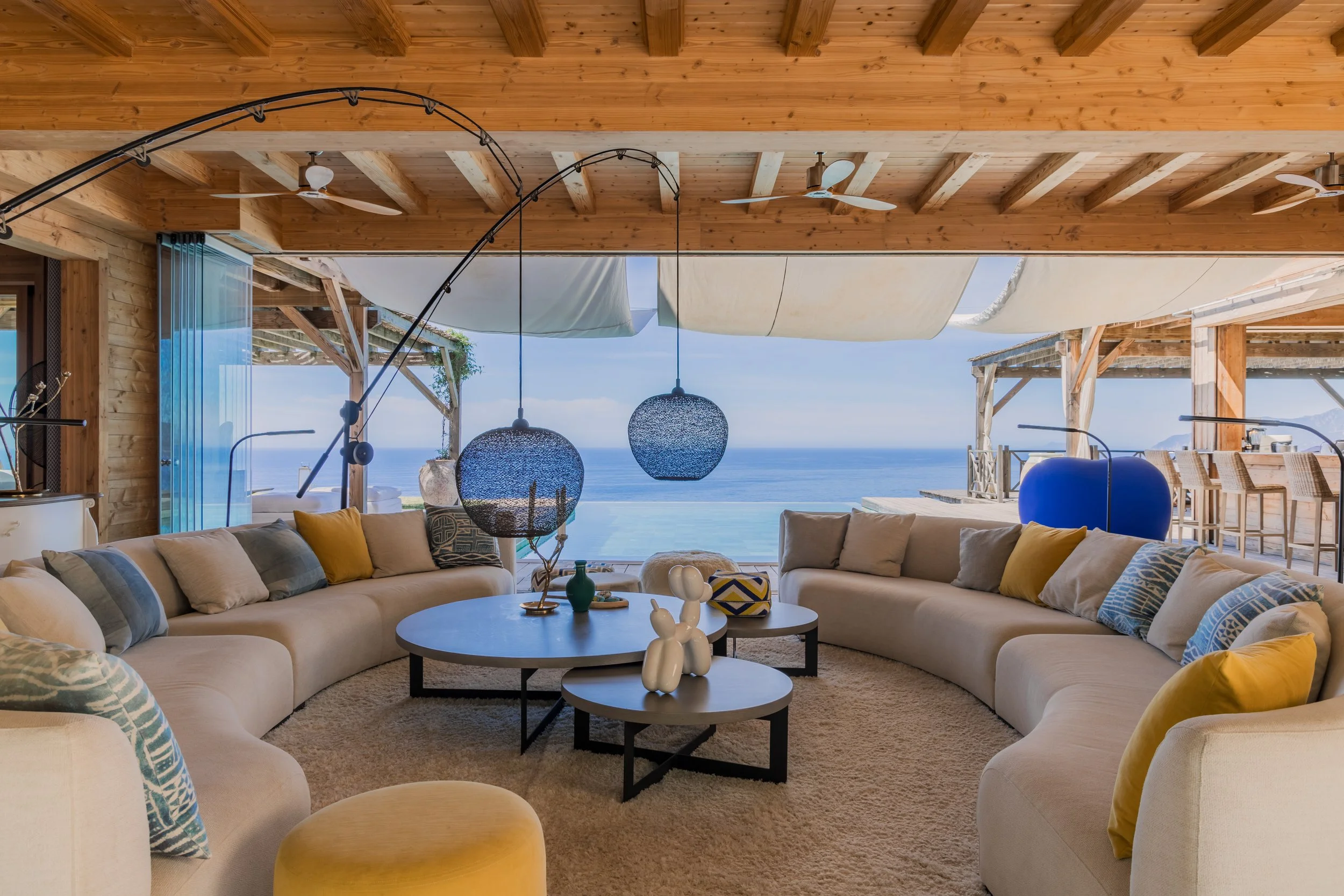 Living room with a large beige sectional sofa with cushions, situated in front of an open wall overlooking the ocean, with a wooden ceiling and hanging pendant lights.