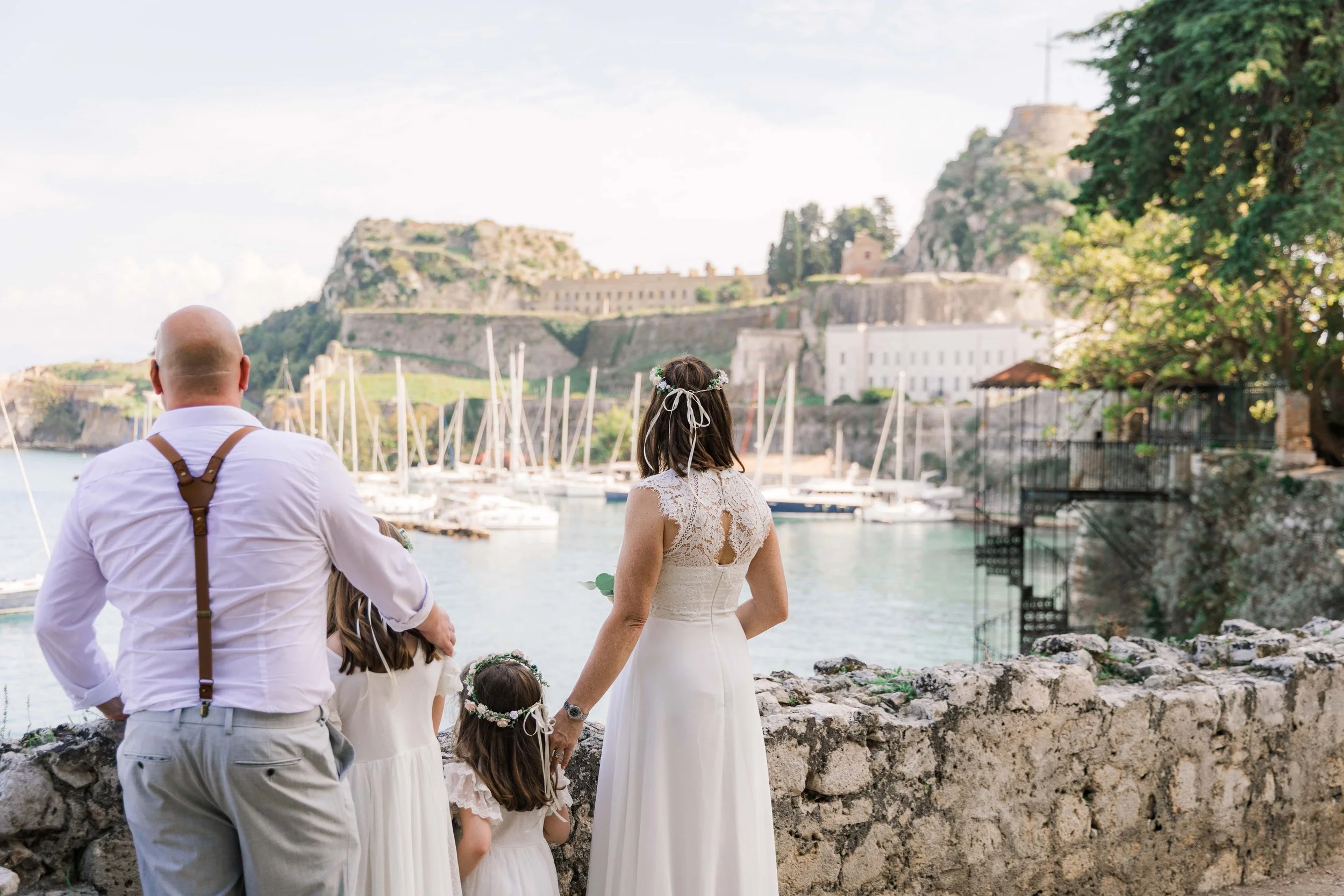 A group of people, including a woman in a white dress and floral crown, two young girls in white dresses with floral crowns, and a bald man in a white shirt with suspenders, standing by a stone wall overlooking a marina with sailboats and a hillside 