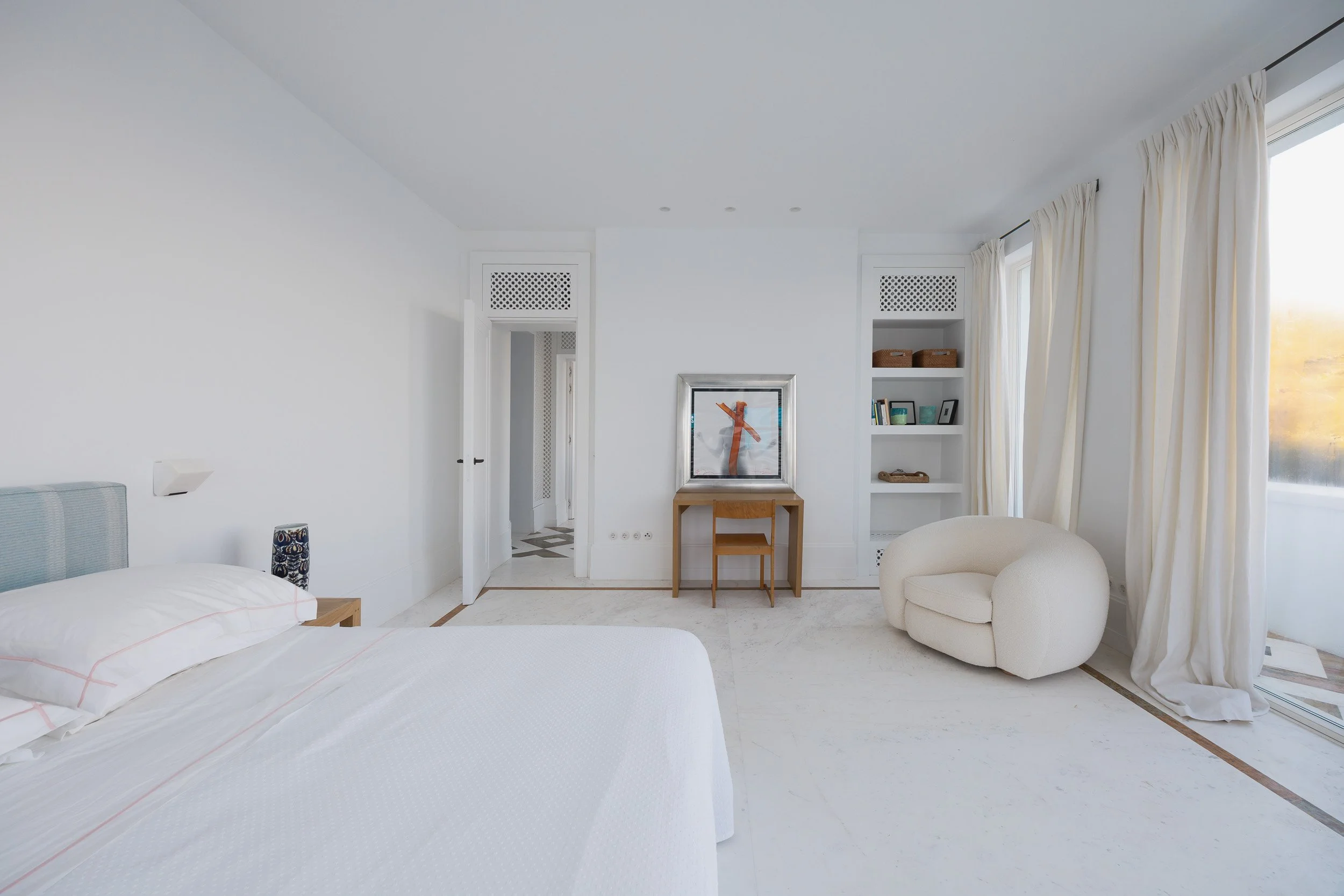 A bright, minimalistic bedroom with a white bed, a small wooden nightstand, a round white armchair, a wooden desk with a painting on it, open shelves with decor, and large windows with cream curtains letting in natural light.