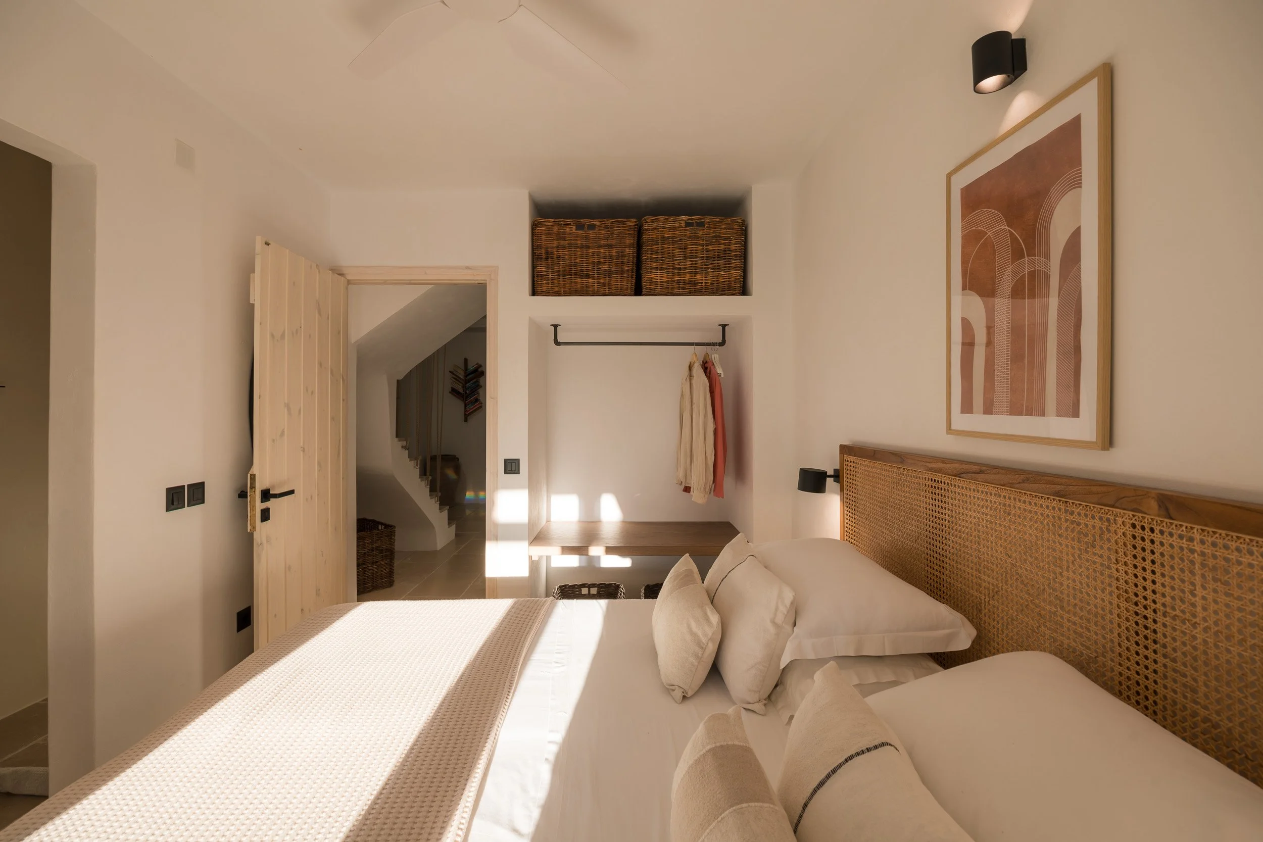 A cozy bedroom with a large bed, white bedding, and a decorative headboard. There is a framed abstract artwork hanging above the bed and a black wall-mounted lamp on each side. The room has a wooden door, a small desk area with hanging clothes, and w