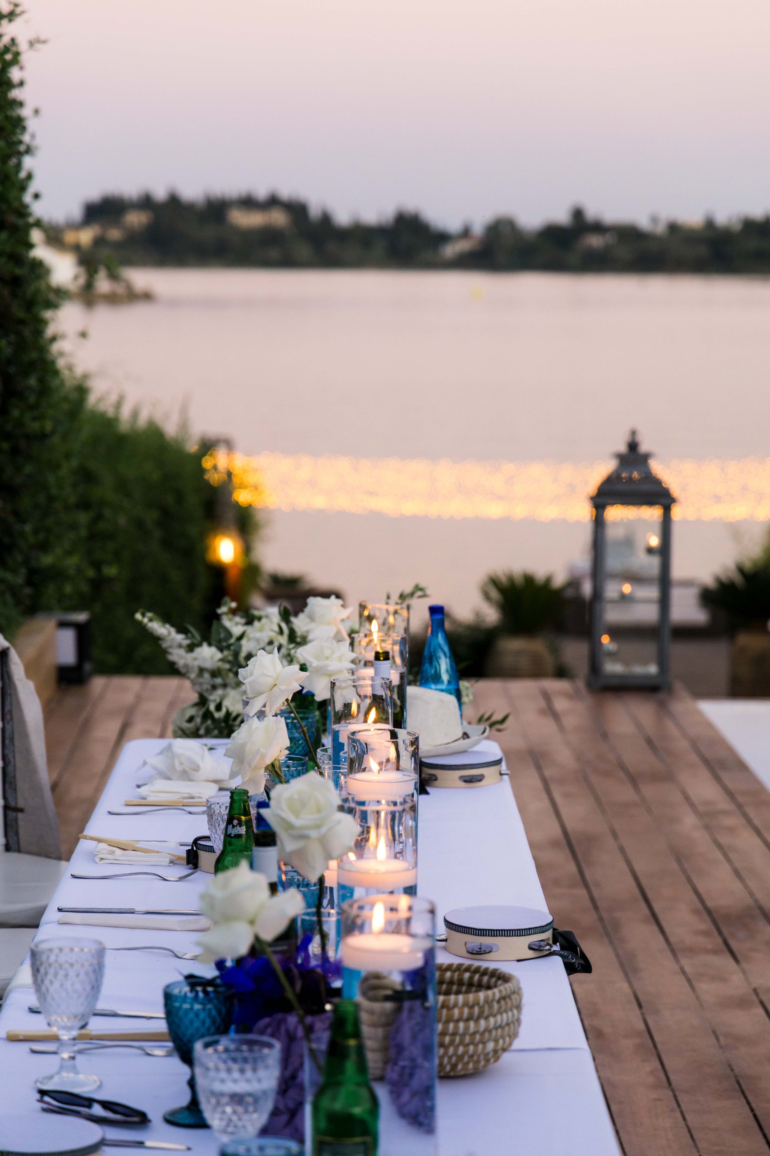 Outdoor dinner table set near water at sunset with white flowers, candles, and blue bottles.