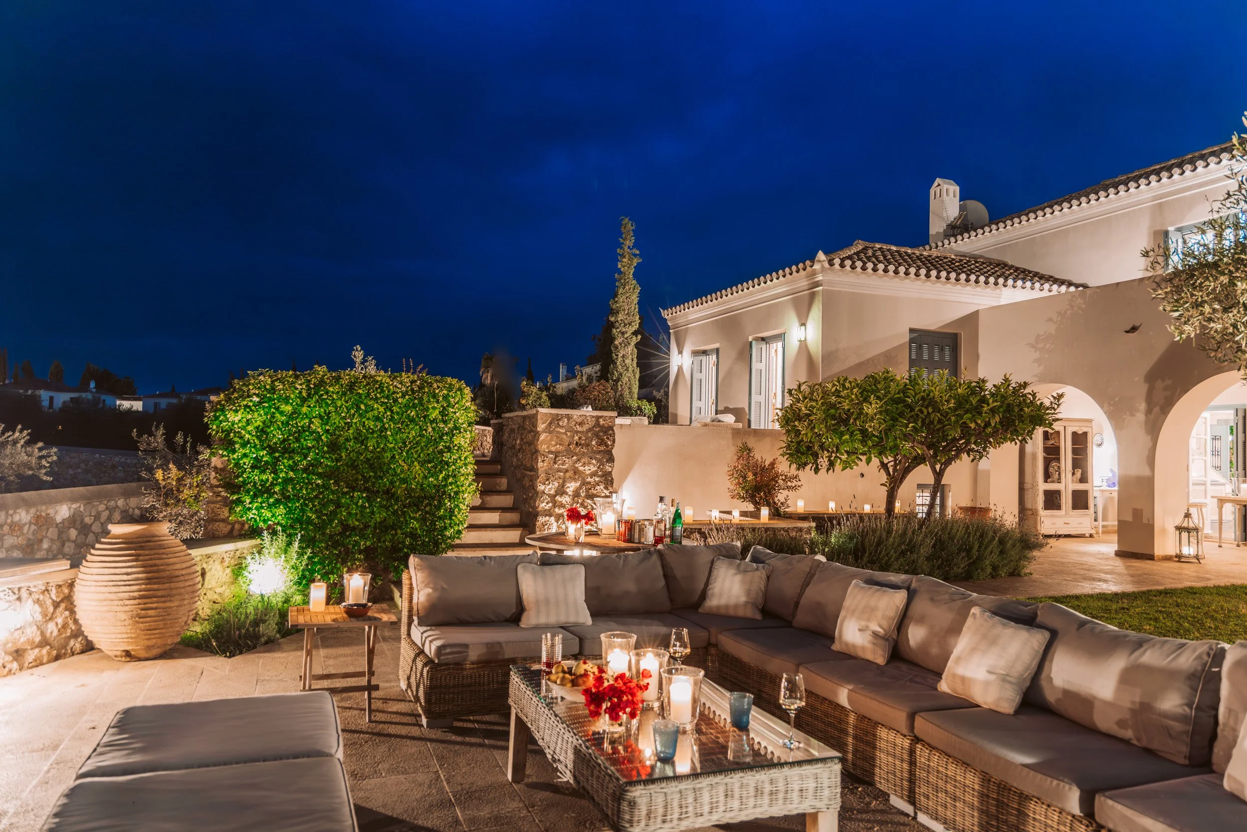 Outdoor patio at night with wicker sectional sofa, candles, and drinks, in front of a white house with arched doorways and shutters, surrounded by trees and greenery, illuminated by outdoor lights.